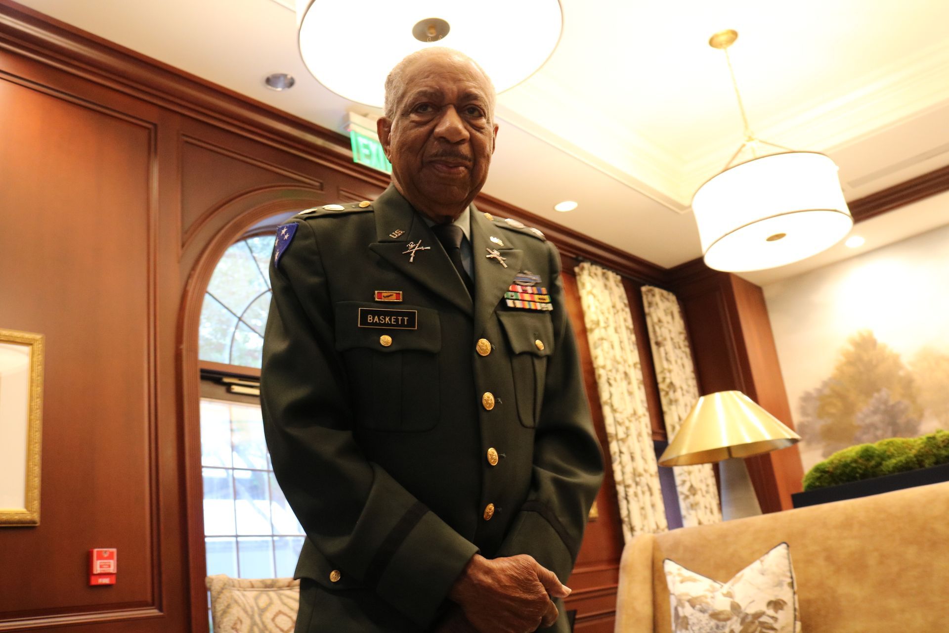 Man in military uniform stands indoors, arms at sides, looking at the viewer.