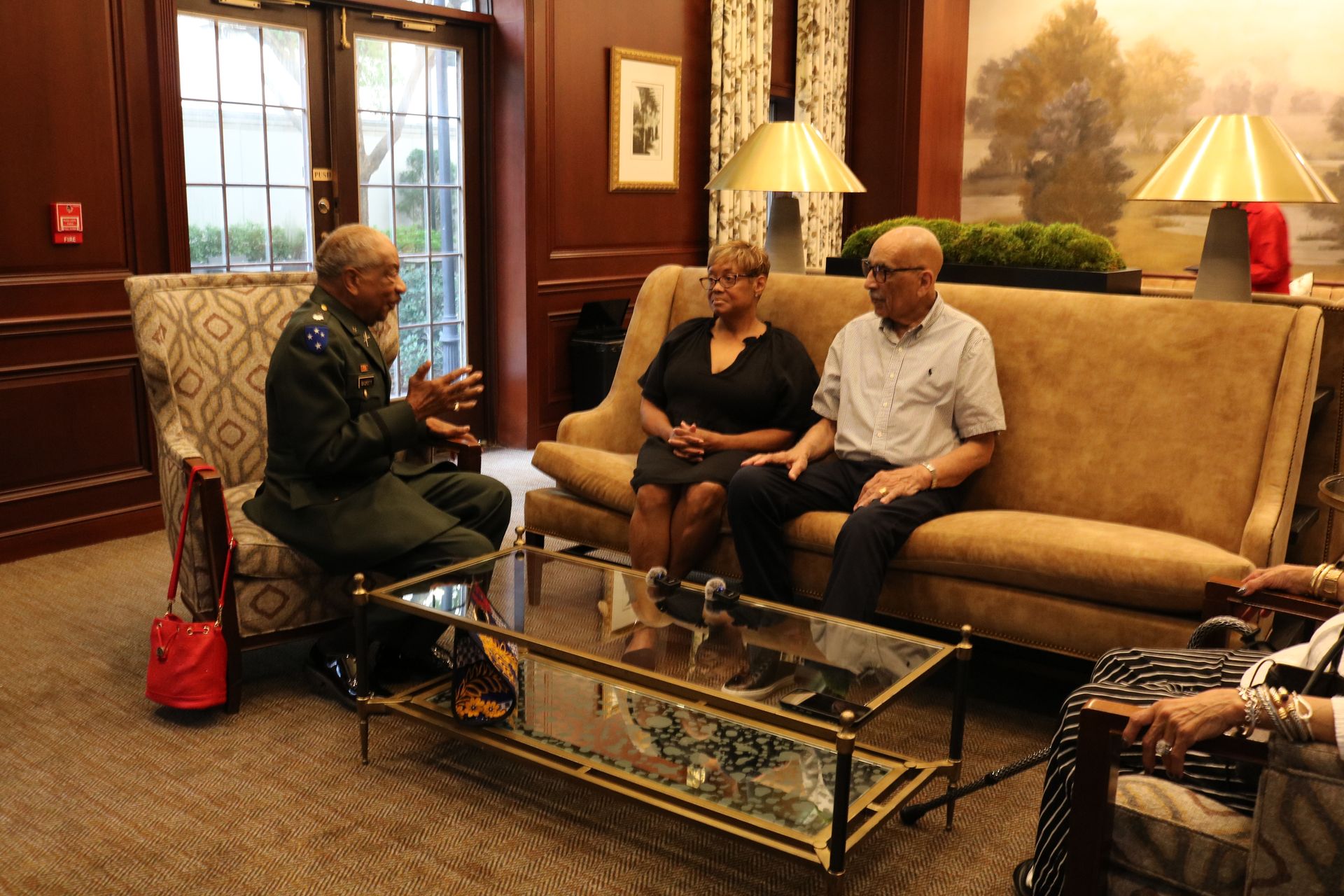 Man in military uniform conversing with a couple seated on a couch in a warmly lit room.