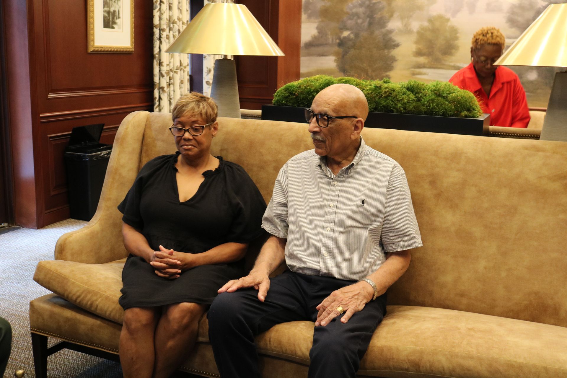 Two people sitting on a sofa in a room. A person in red is in the background.