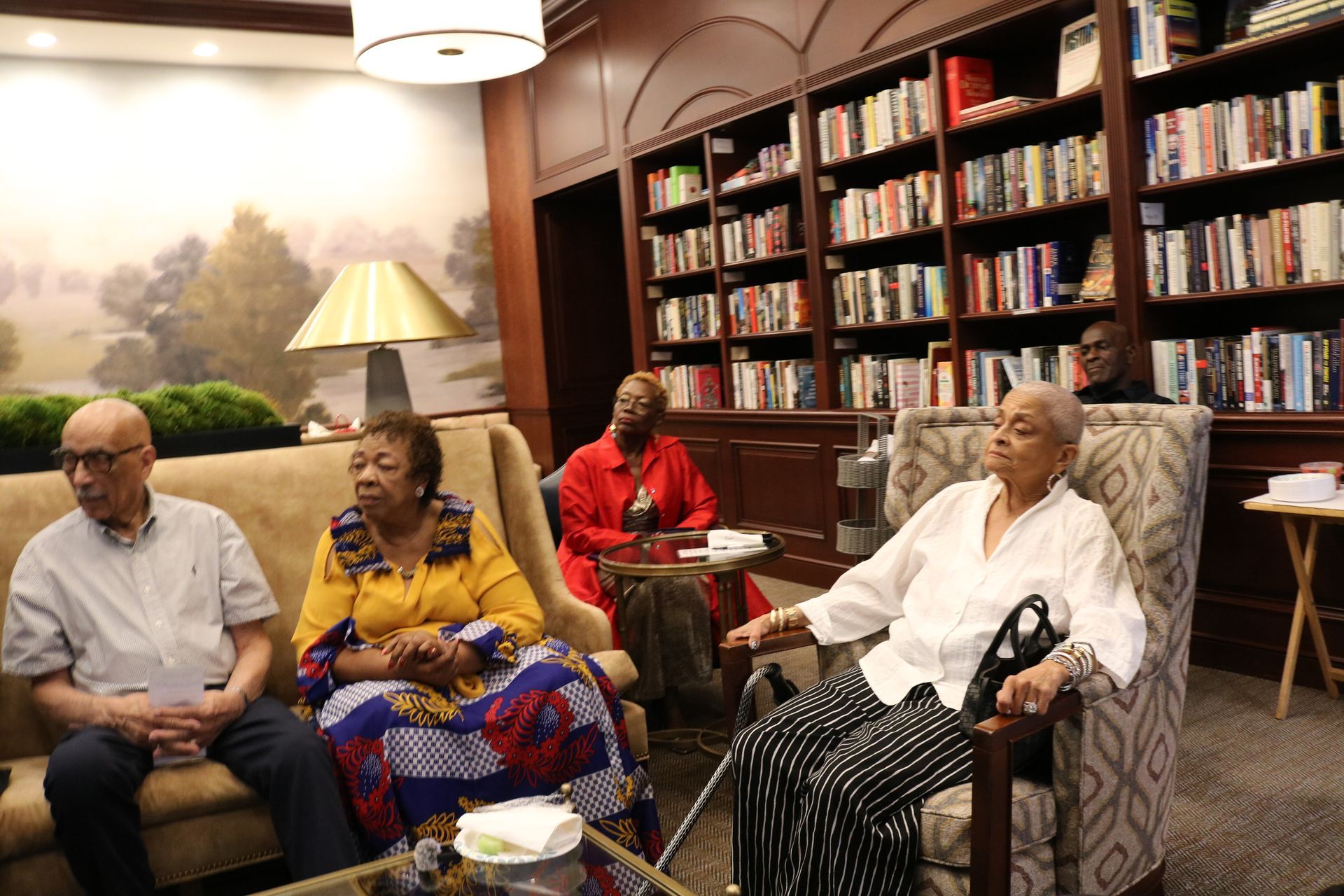Group of people in a library, seated on a couch and chairs, listening. Bookshelves and a lamp are in the background.