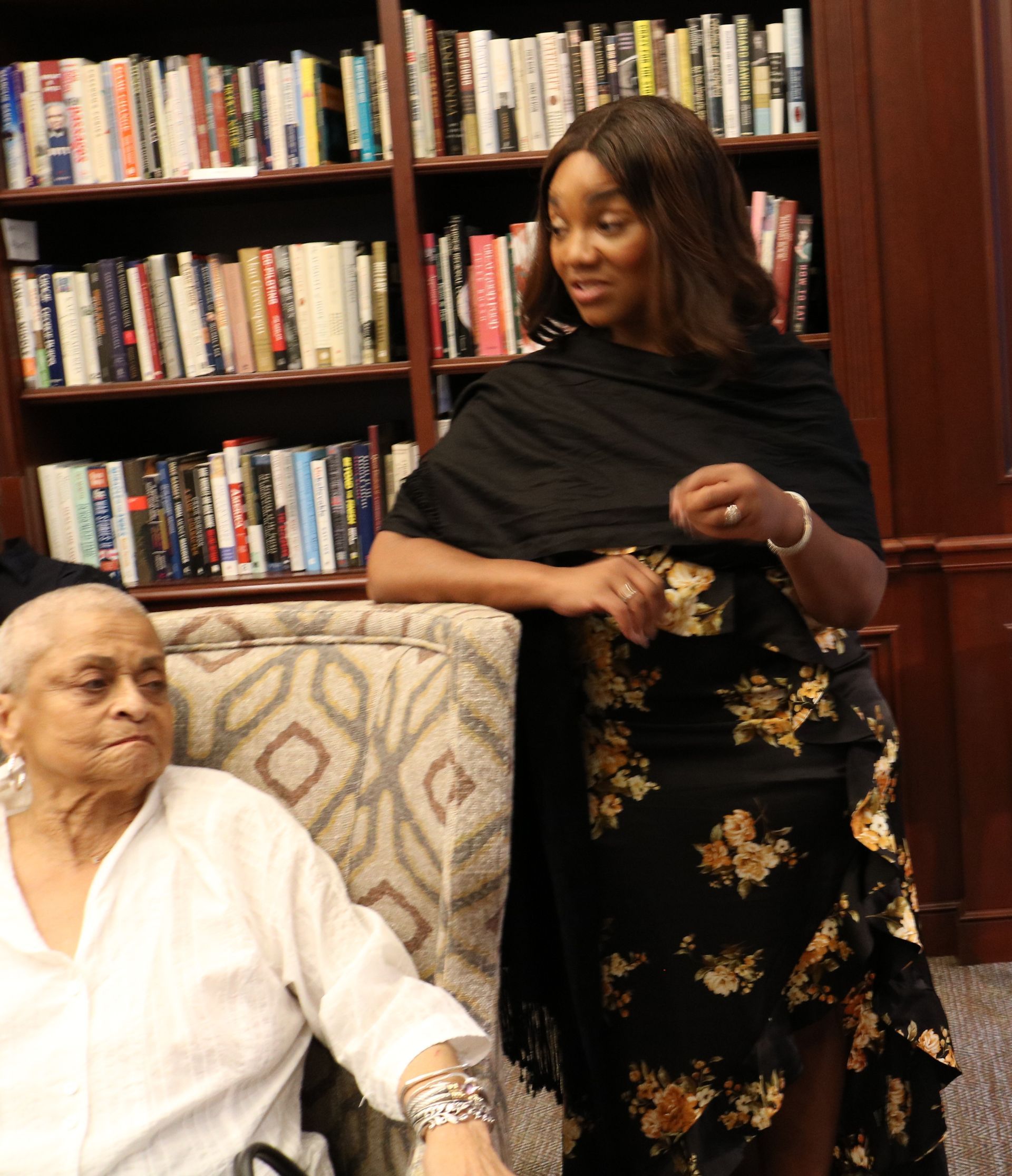 Woman in floral dress and shawl stands near an older person seated in front of a bookshelf.