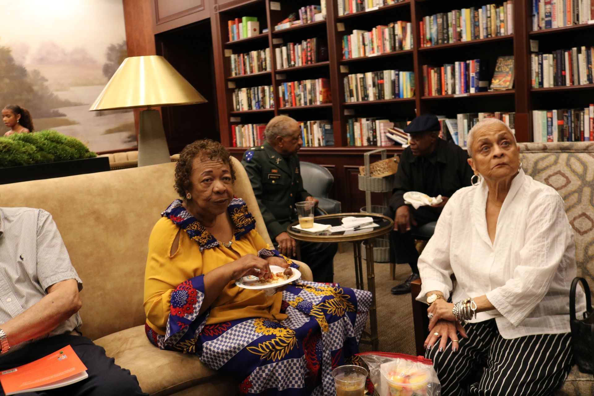 People seated in a library, one eating from a plate, others conversing. Bookshelves in the background.