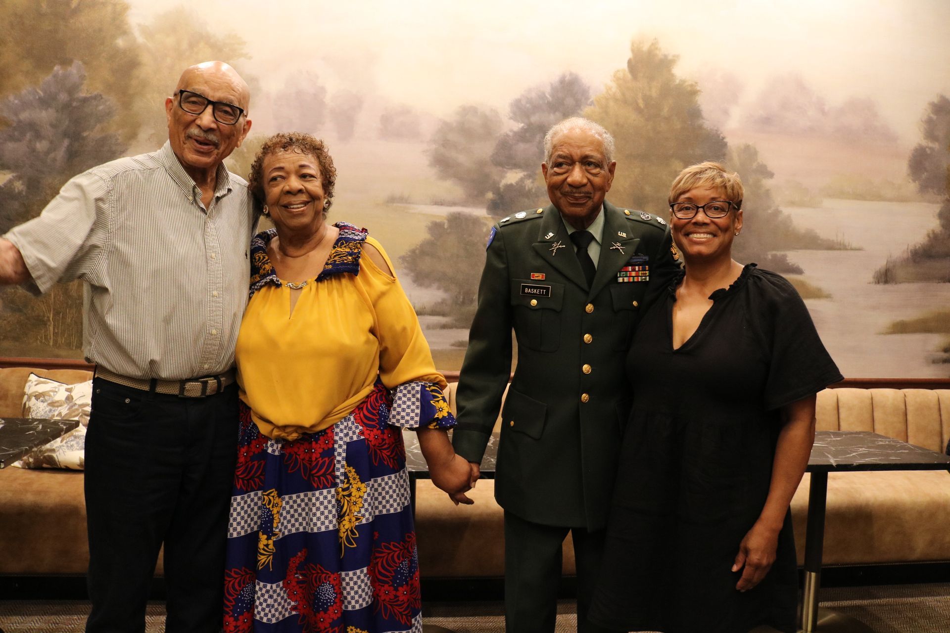 Four people smiling, holding hands, in front of a painted wall. Man in military uniform.