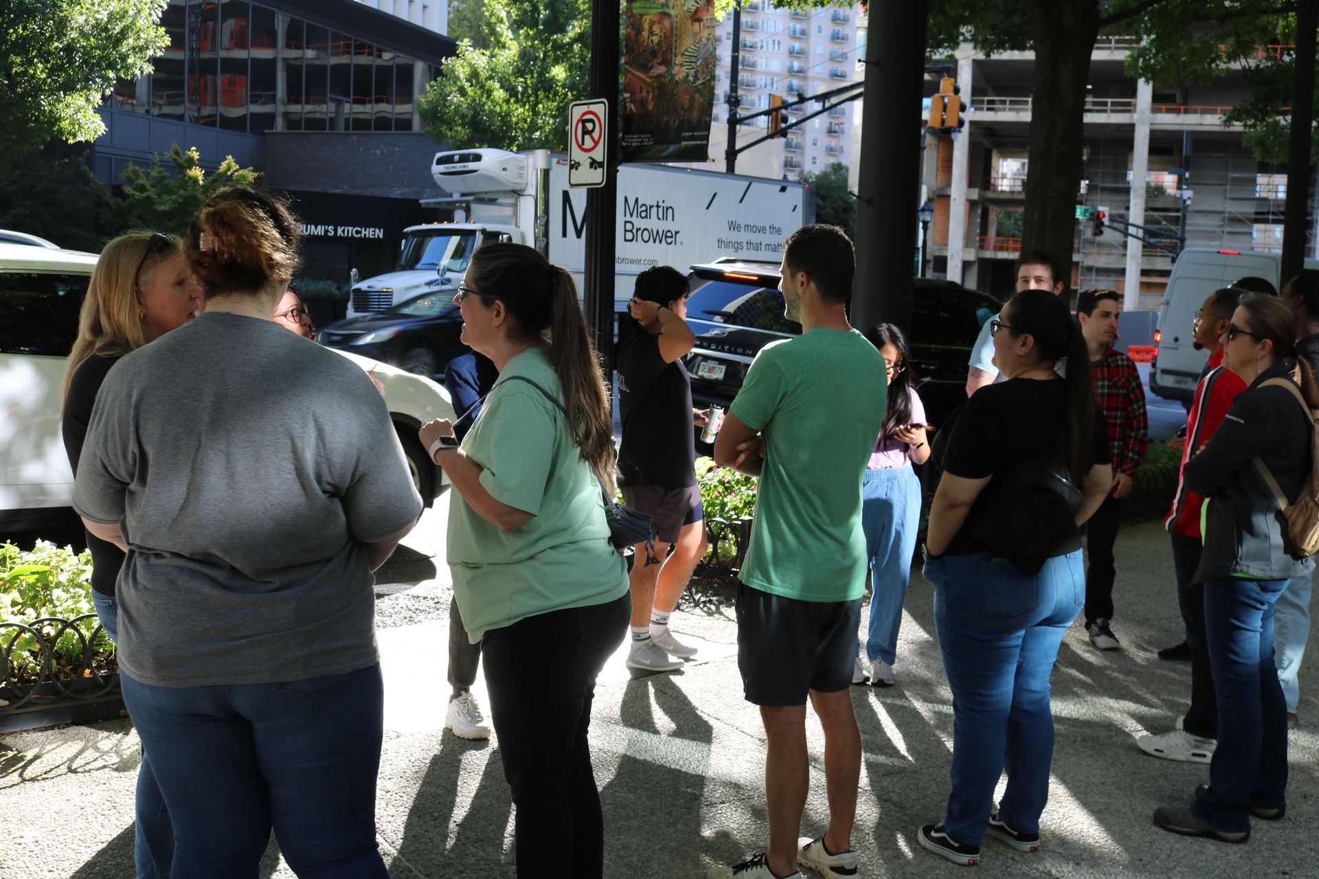 Group of people standing outside, possibly on a city sidewalk. Some are talking, others looking around.