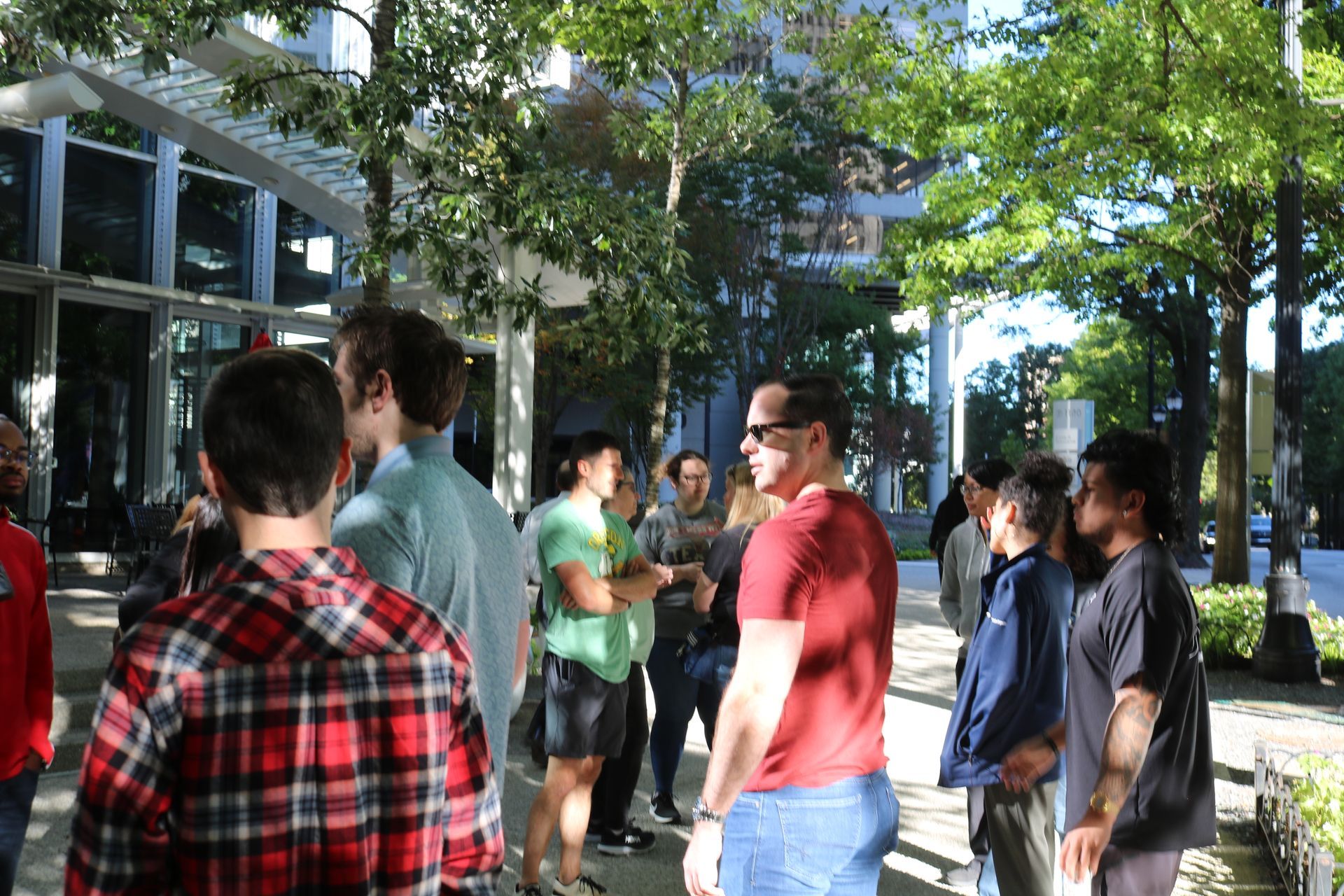 Group of people socializing outside a building with glass windows, under trees. Sunlight illuminates the scene.