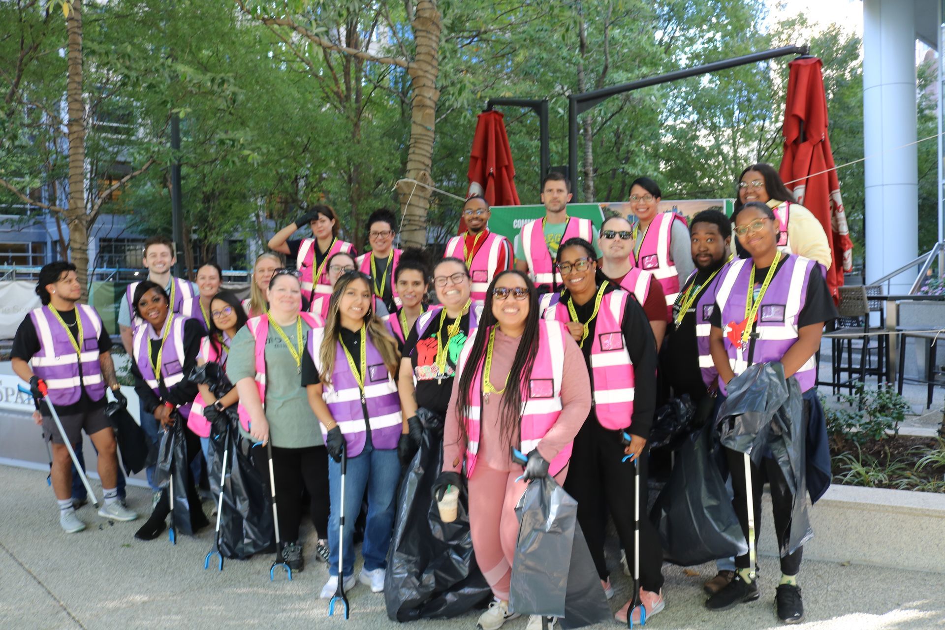 Group of people in pink/purple vests holding trash bags and cleaning outdoors.
