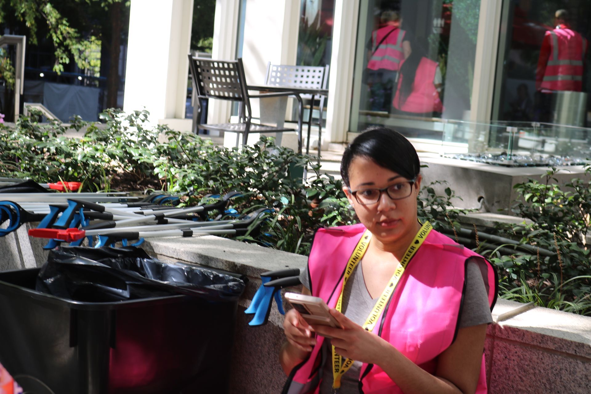 Woman wearing pink and yellow vest, looking at phone. Outdoor setting with debris and building in the background.