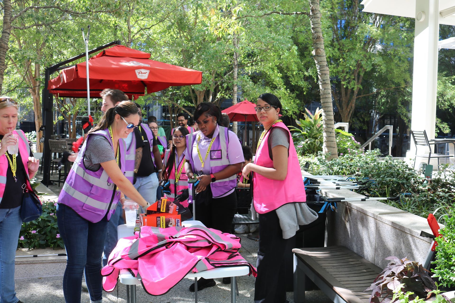 People in pink vests gather near a table under red umbrellas, likely preparing or distributing something outdoors.