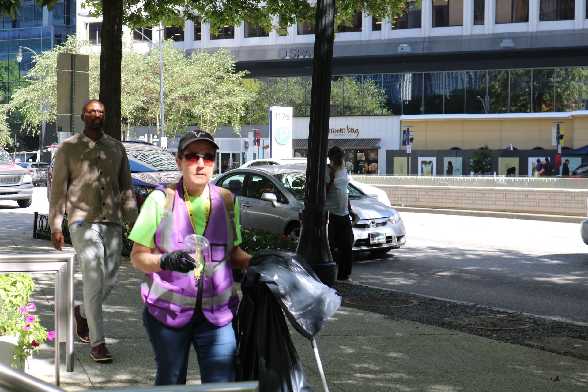 Woman in a purple vest picks up trash on a city sidewalk. A man and cars are in the background.
