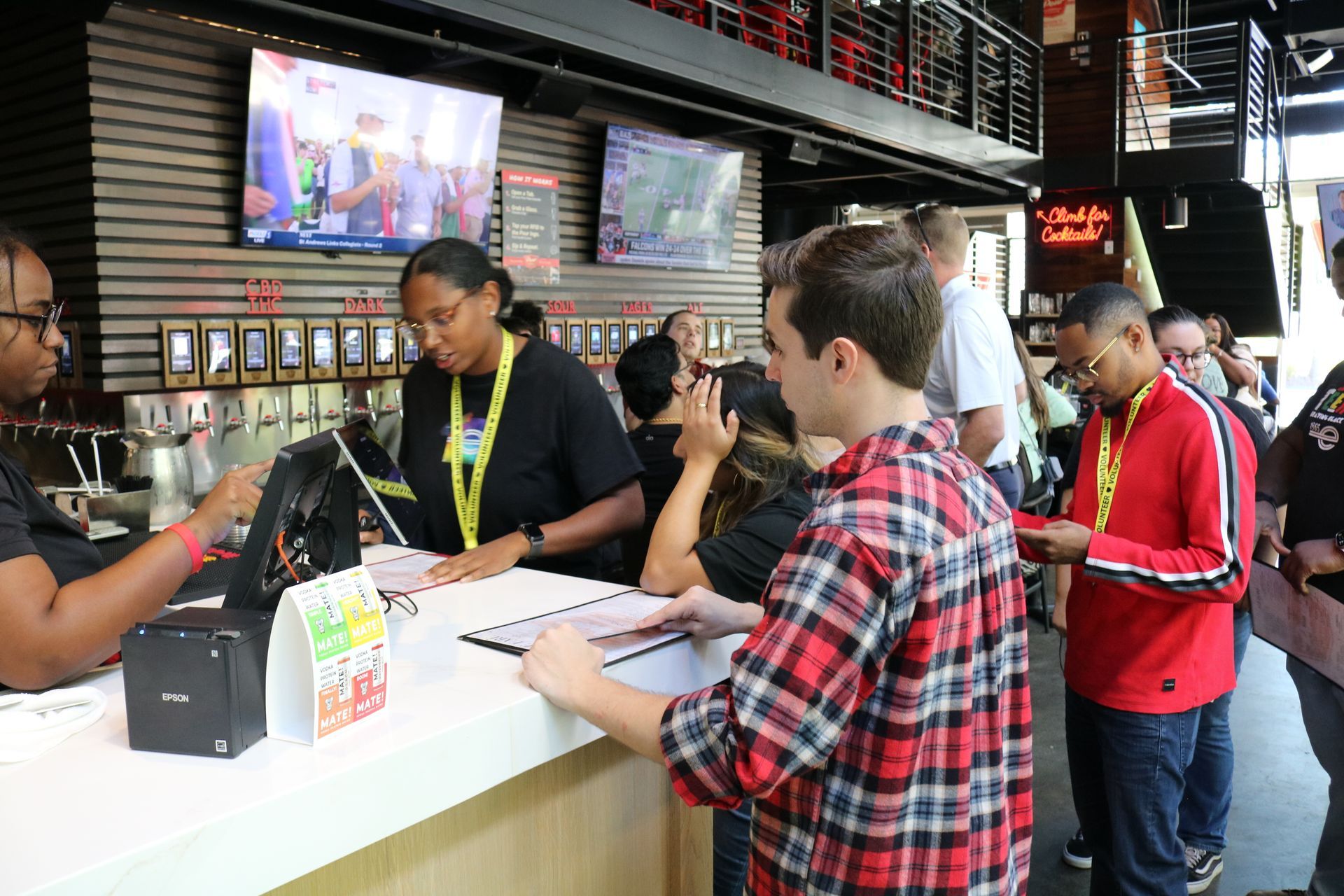 People ordering at a bar, some wearing lanyards. A server is taking an order. TVs are mounted on the wall.