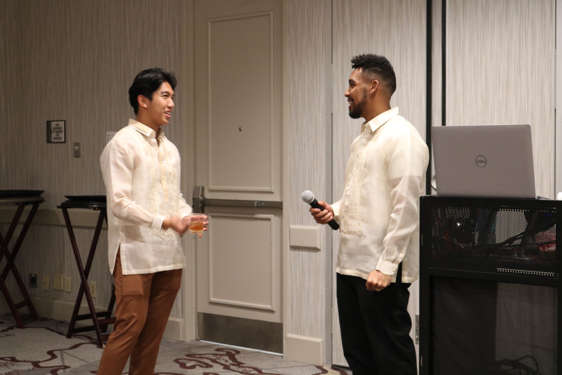 Two men in formal shirts, one holding a mic, the other a drink, speaking near a laptop.