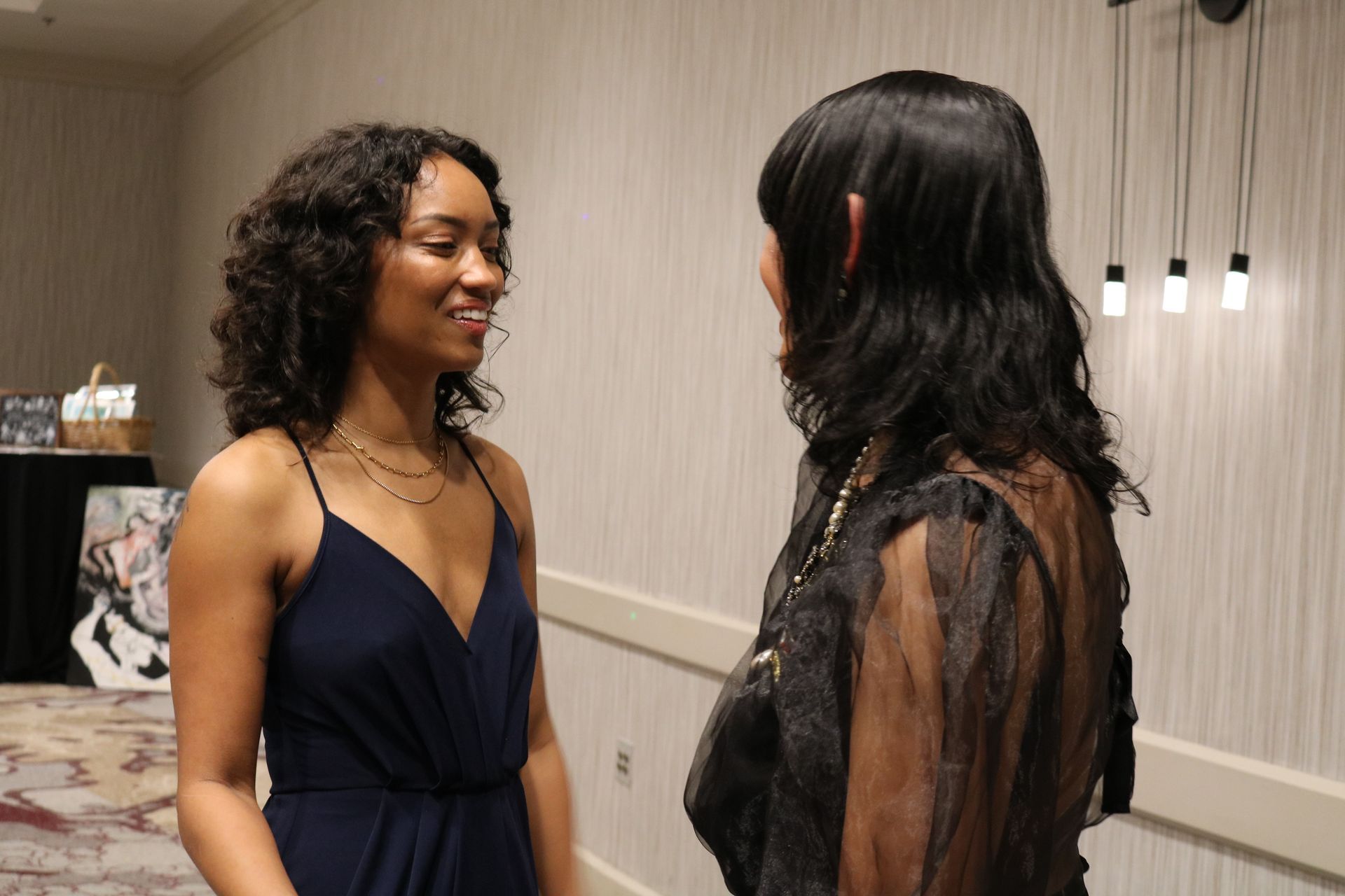 Two women converse in a well-lit hallway. The woman on the left smiles in a navy dress, the other in black lace.