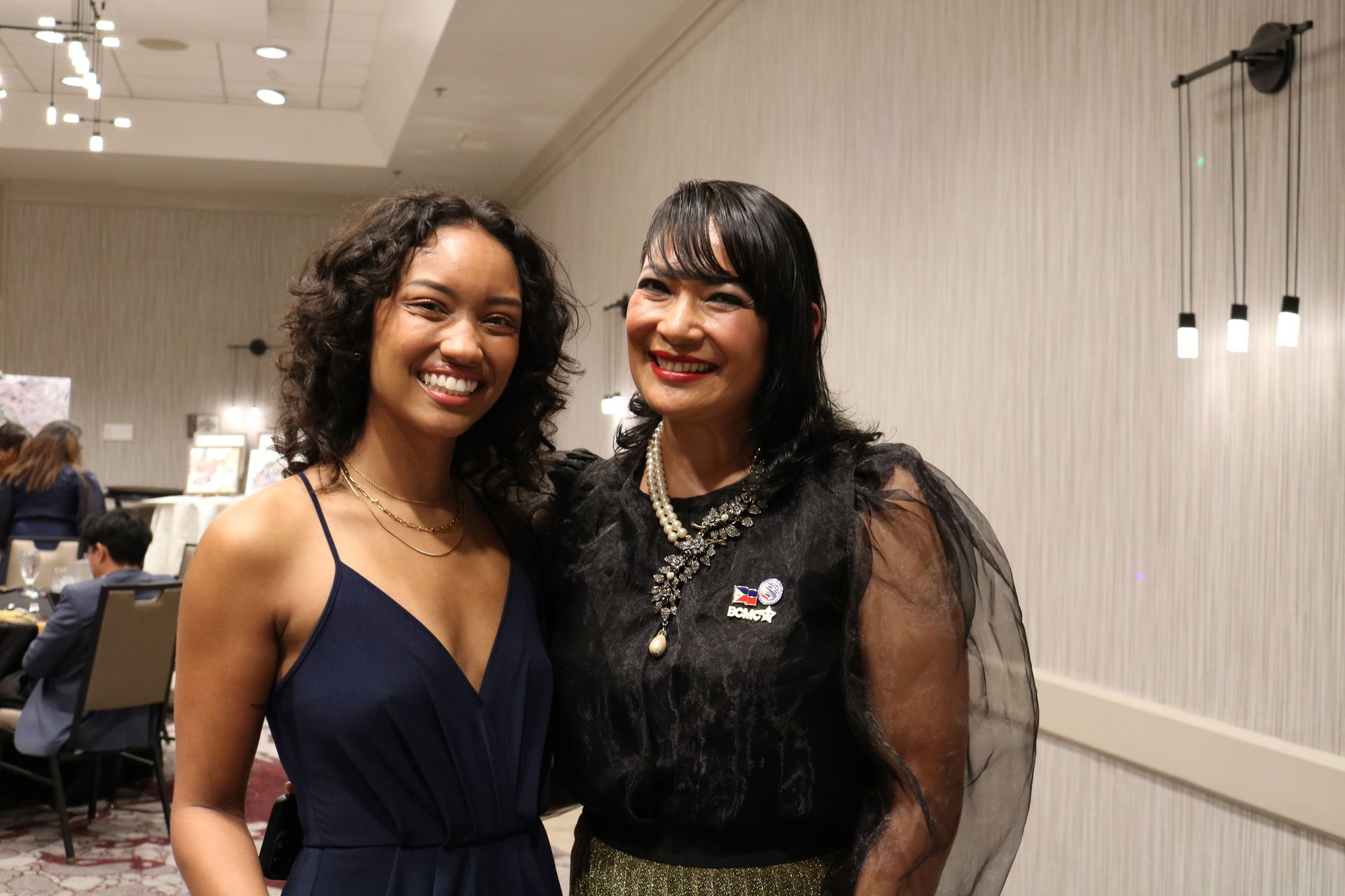 Two smiling women pose indoors; one in a navy dress, the other in black, ornate attire.