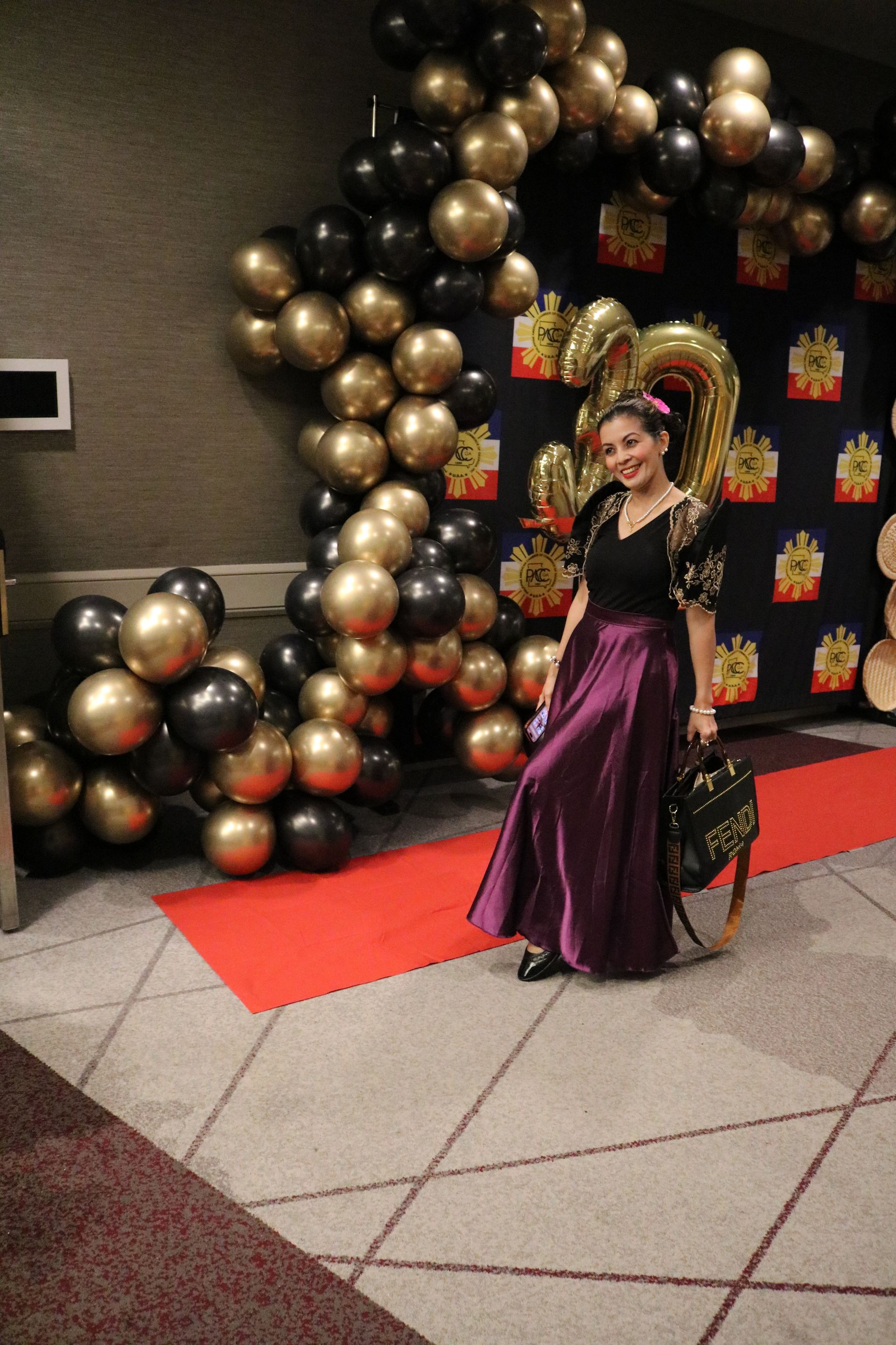 Woman in purple dress poses on red carpet by gold and black balloon arch.
