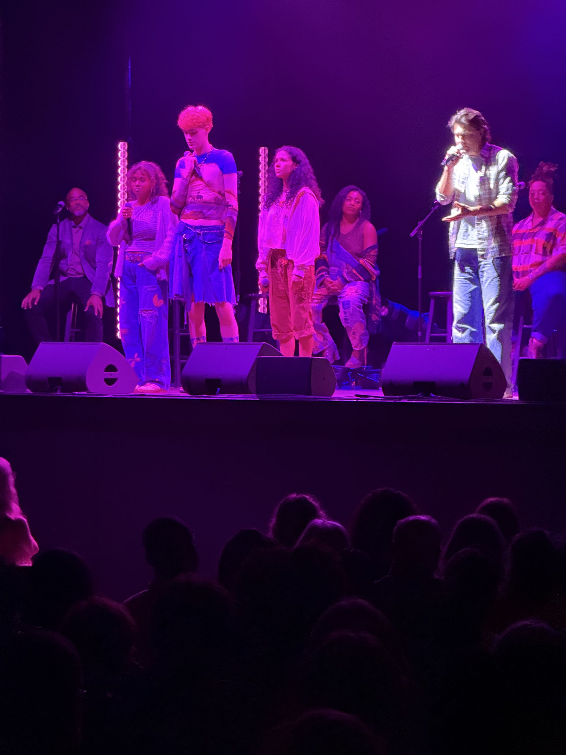 A group of singers on stage under purple lights perform to an audience.