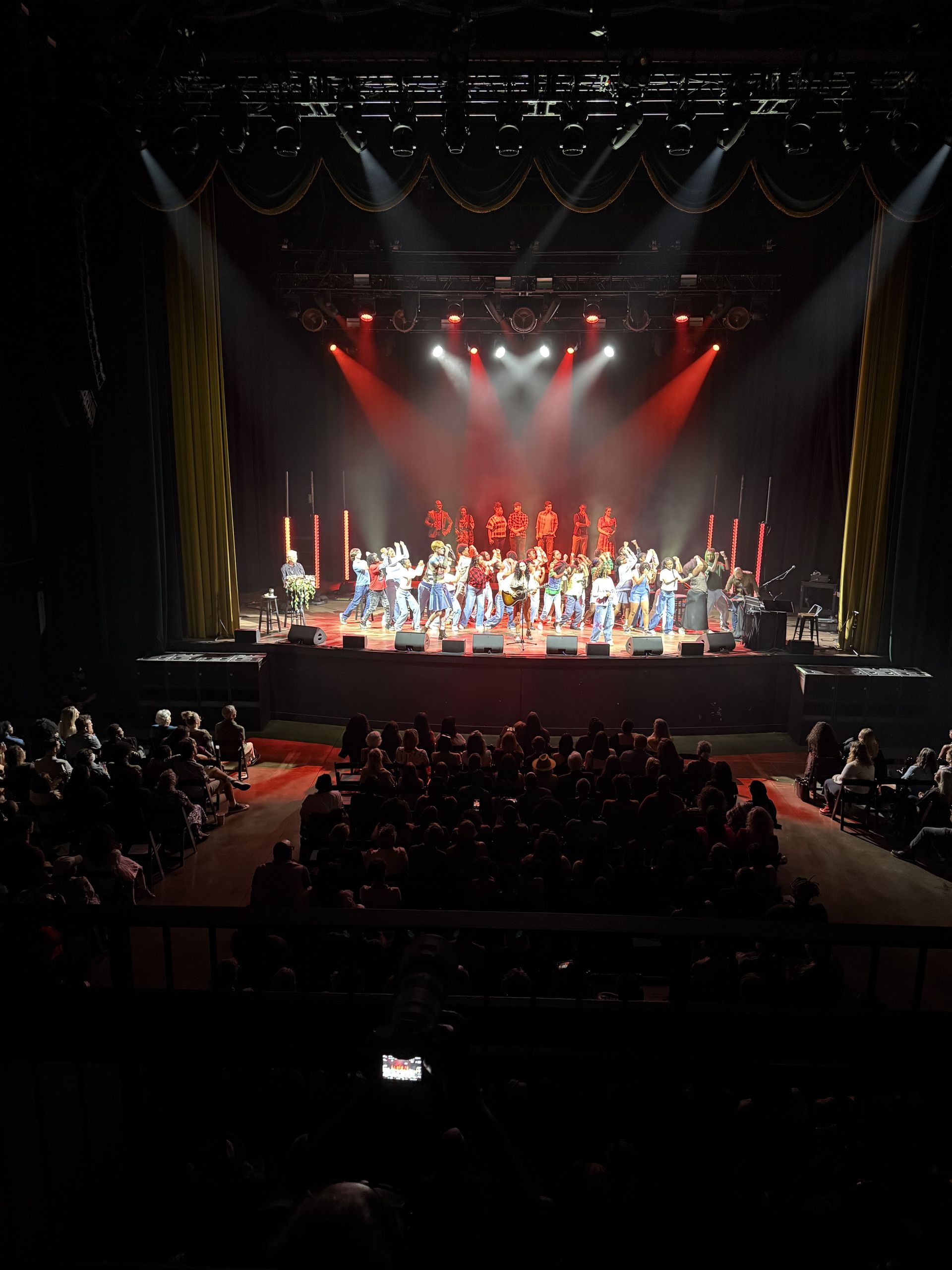 Audience watches a large group performing on a stage lit with red and white lights.