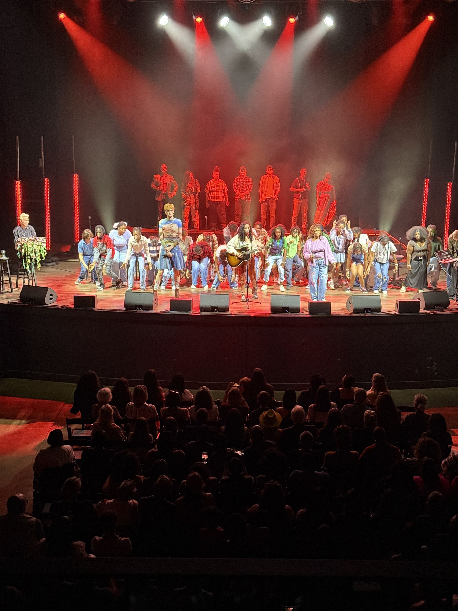 Concert stage lit with red lights. Large group of musicians performing; audience in the foreground.