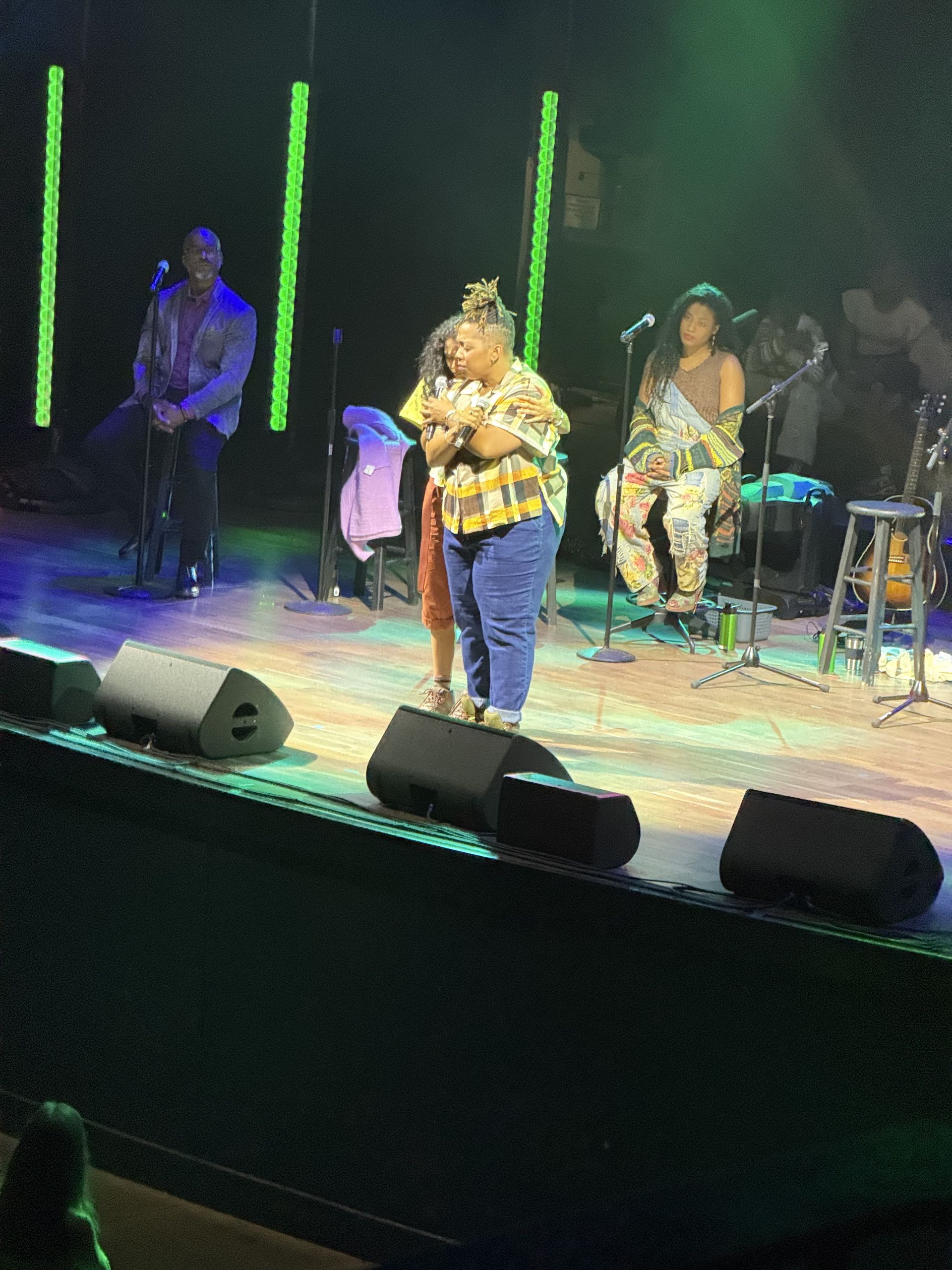 Woman onstage performing at a venue. Two others seated behind her, green lights.