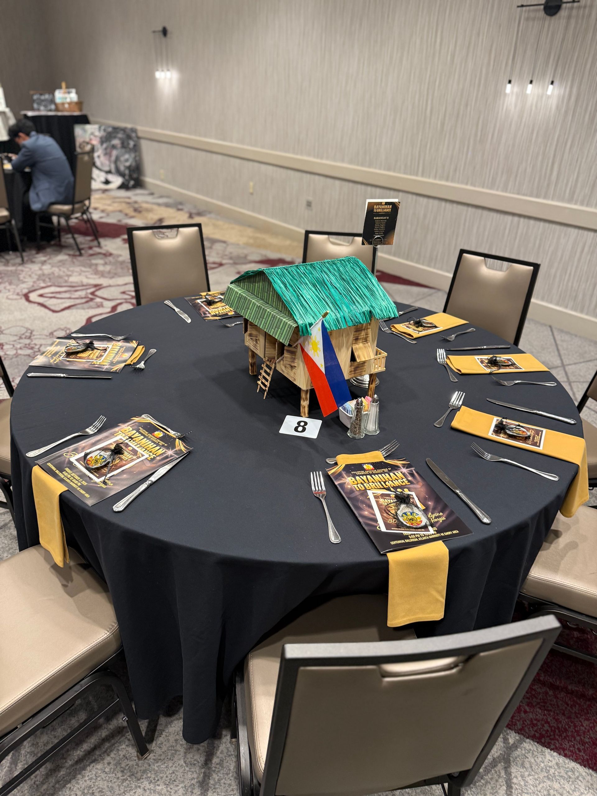 Round table set for a formal dinner with a centerpiece of a miniature house and Philippine flag.
