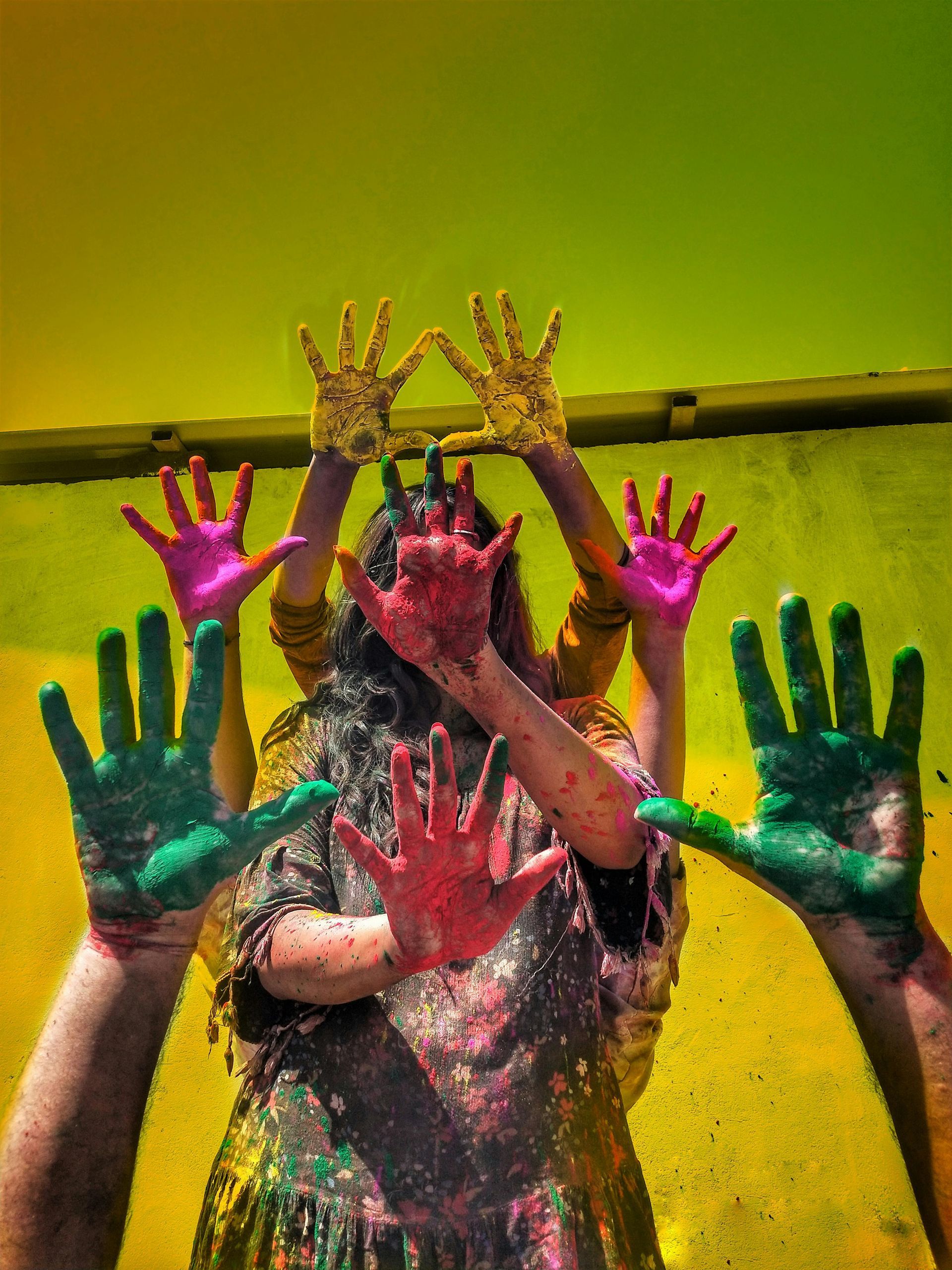 Hands covered in colorful paint are raised toward the camera against a yellow and green background.