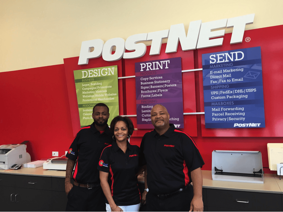 Three people in PostNet uniforms stand in front of a PostNet sign, red wall, various service panels.