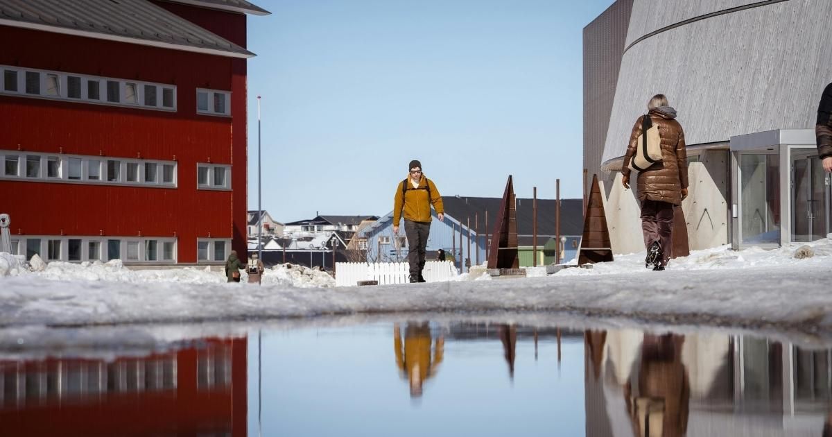 Man walks past a red building and a cylindrical structure; snowy, sunny outdoors.