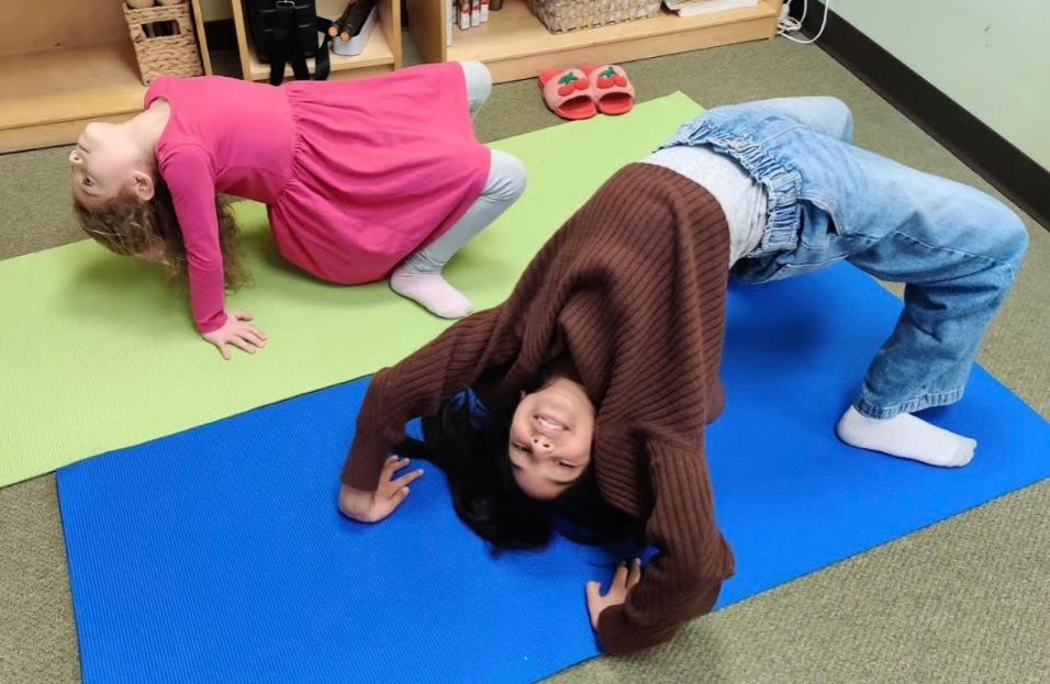 Two children on yoga mats attempting the bridge pose, and smiling.