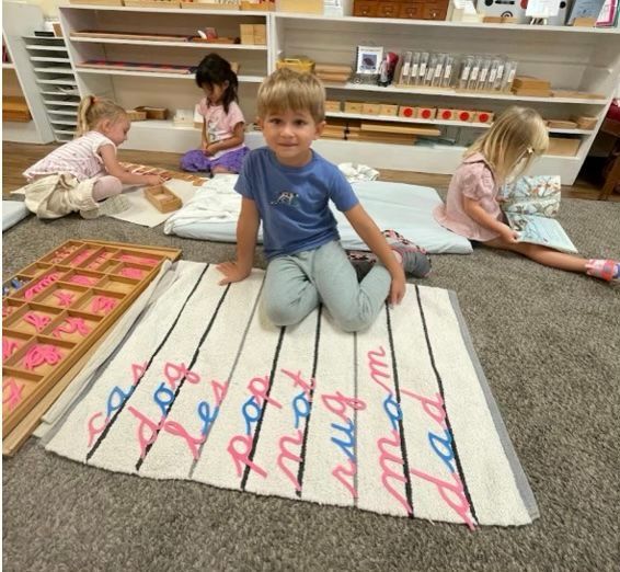 A young child sits on the floor by a rug that has short words spelled out with pink and blue letters