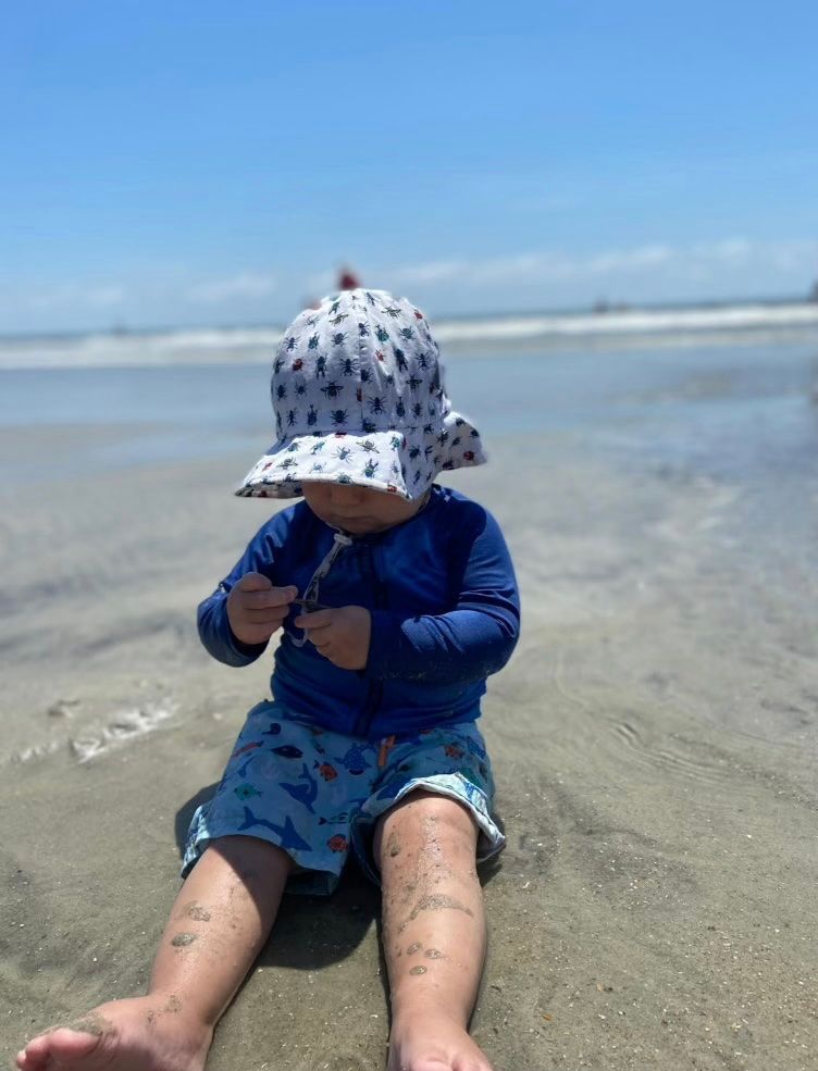 A young child wearing swimming gear sits on a beach with the ocean behind him.
