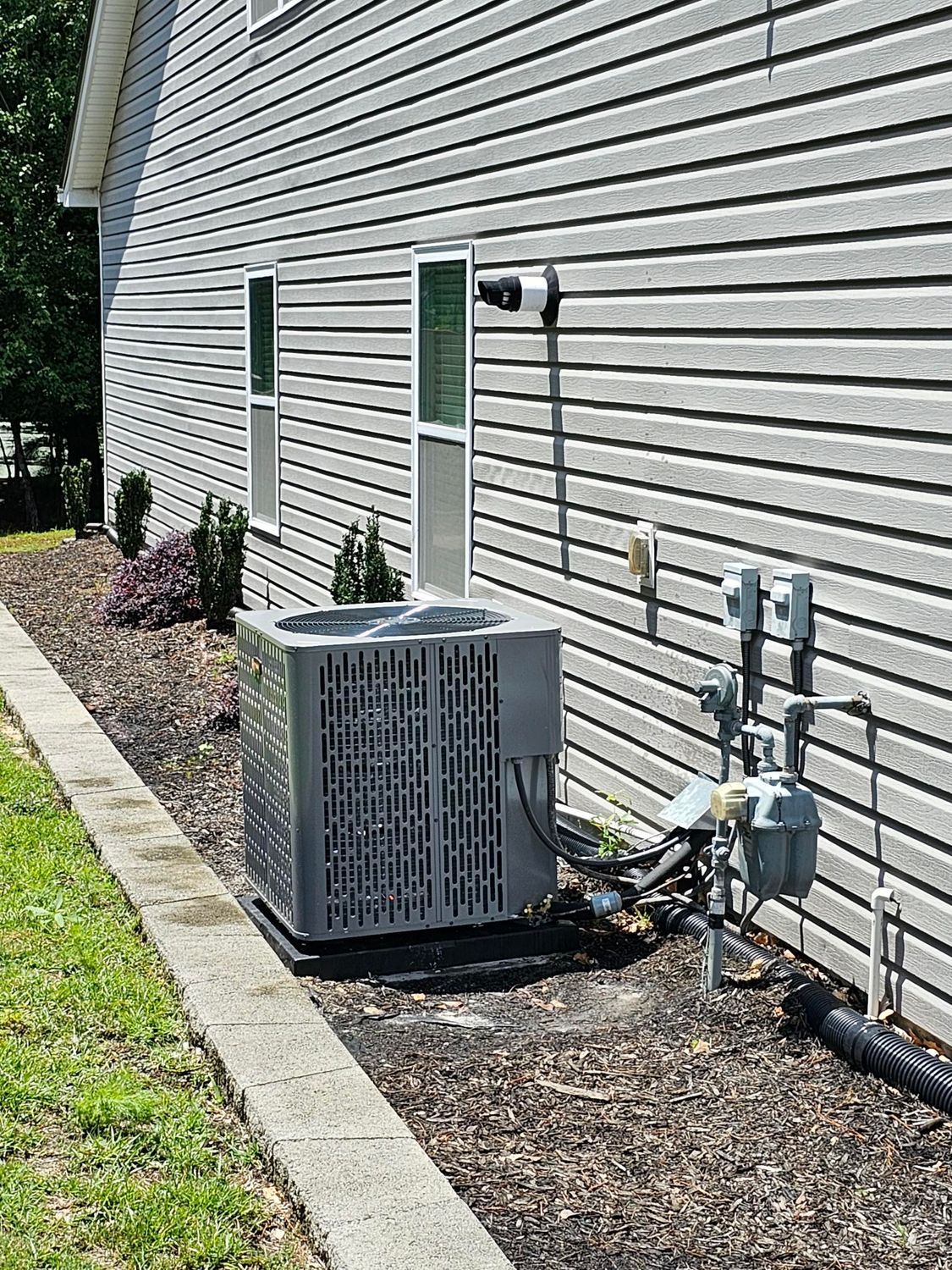 Air conditioning unit next to a house with gray siding. Gas meter and landscaping visible.