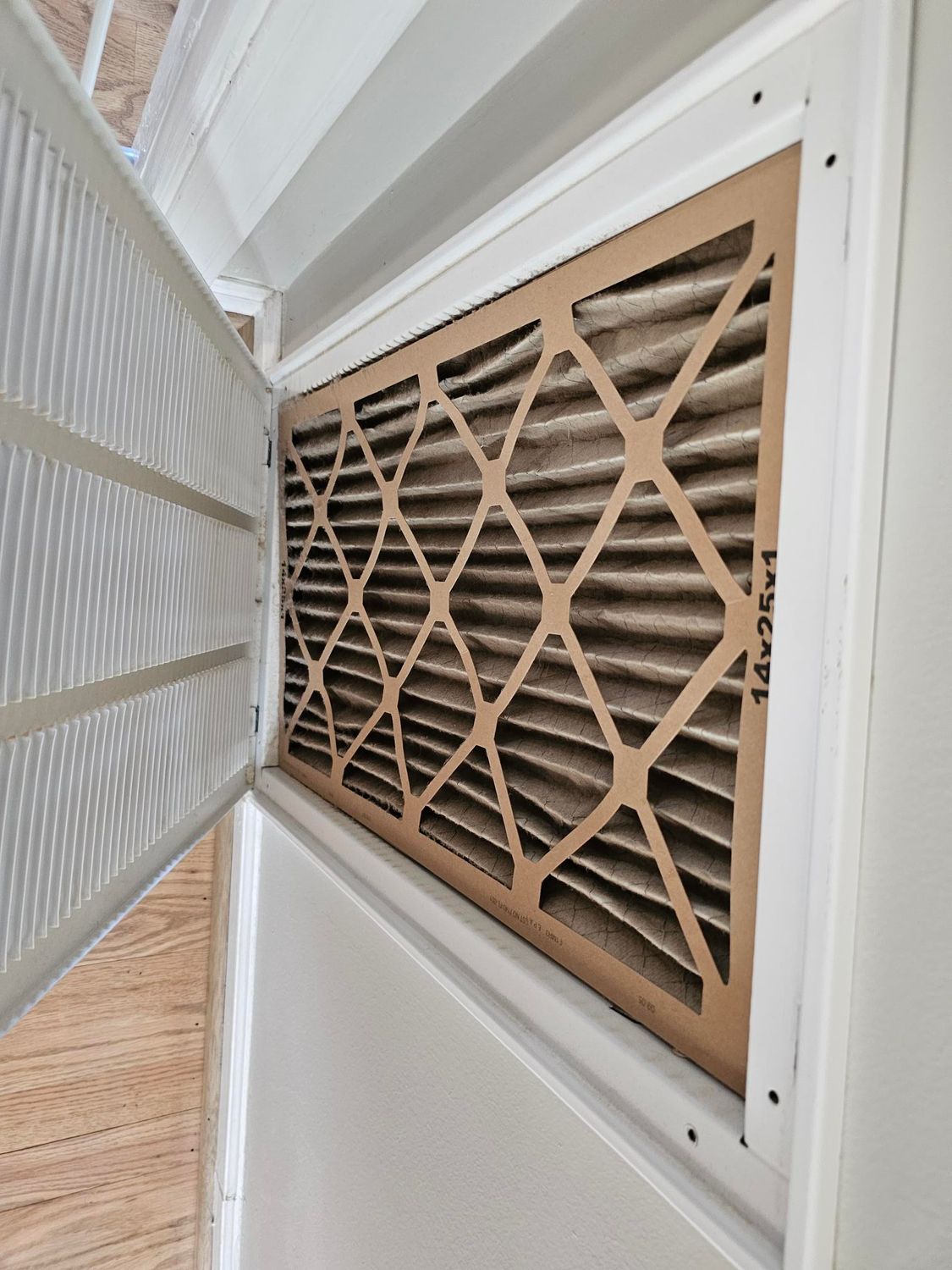A dirty air filter inside a white metal housing. The filter is brown, accordion-style, and needs replacement.
