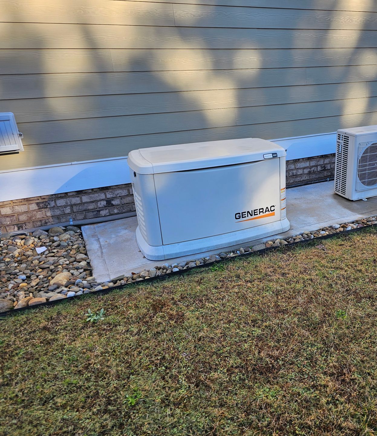 A white Generac generator installed next to a brick wall and air conditioning unit on a concrete pad.