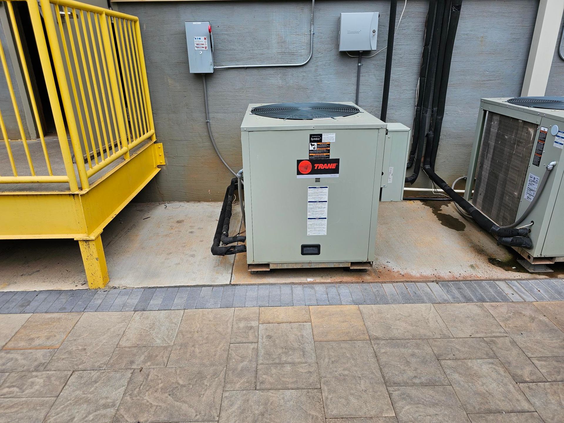 Outdoor HVAC unit next to a yellow railing and a gray wall with electrical boxes.