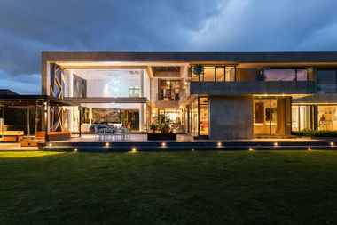 Modern house at dusk with illuminated interior and exterior lights, green lawn, cloudy sky.