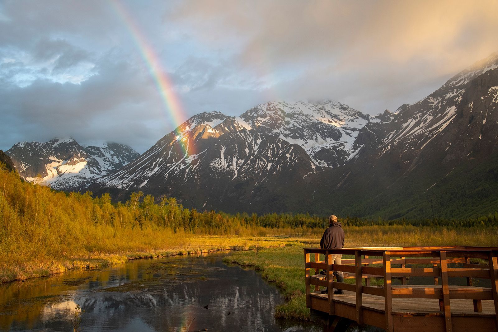 A man is standing on a bridge over a river with a rainbow in the background.