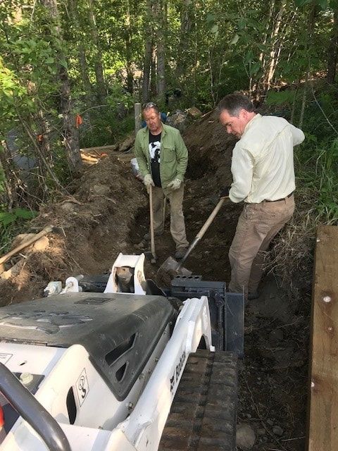 Two men are digging in the dirt next to a bobcat tractor.