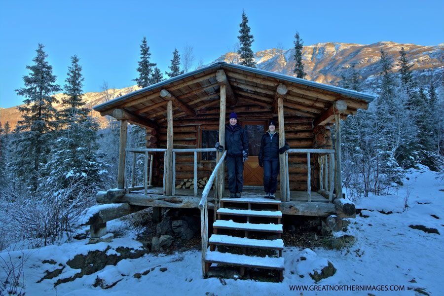 Two people are standing in front of a log cabin in the snow.