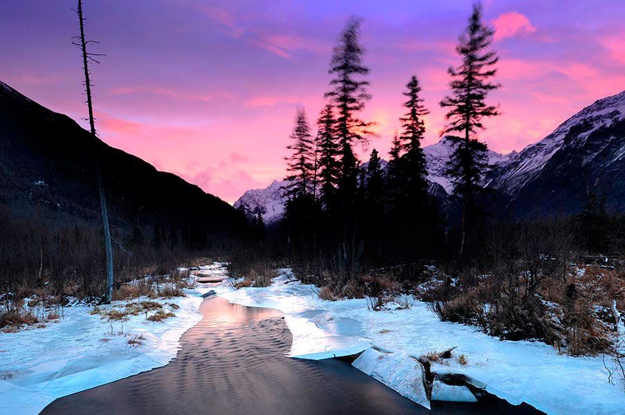 A river running through a snowy forest with mountains in the background