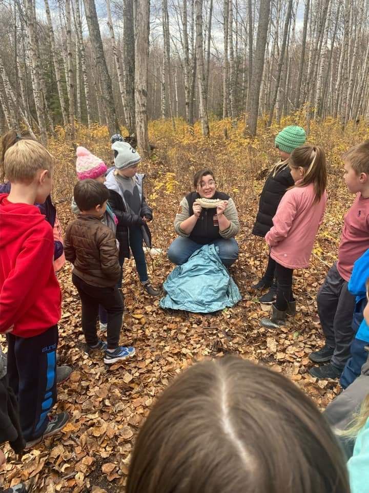 A group of children are standing around a man in the woods.