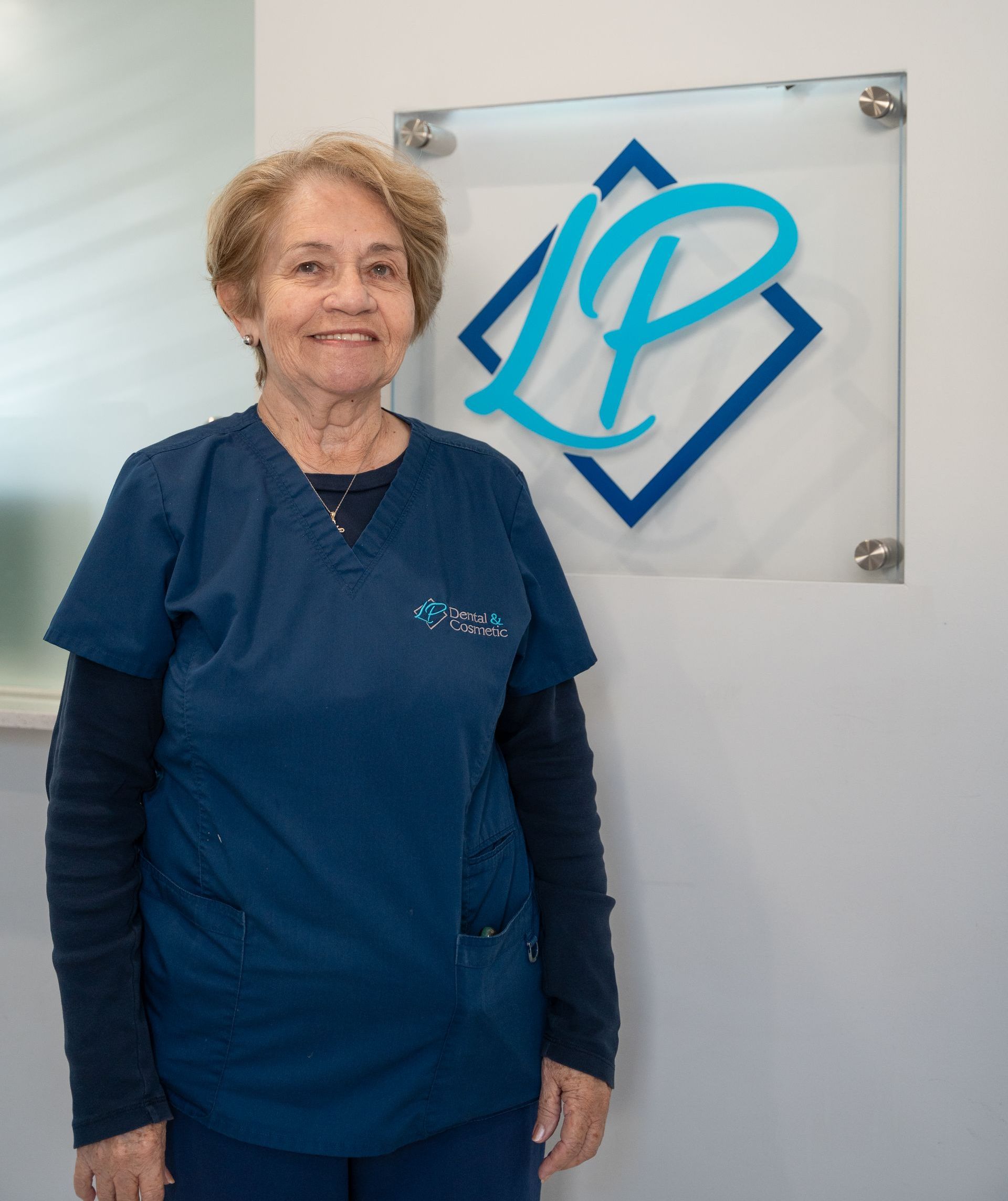 A woman wearing glasses and a blue scrub top is smiling.