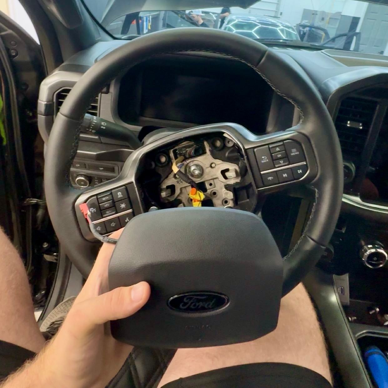 Person holding a Ford airbag cover in a vehicle's interior with the steering wheel exposed.