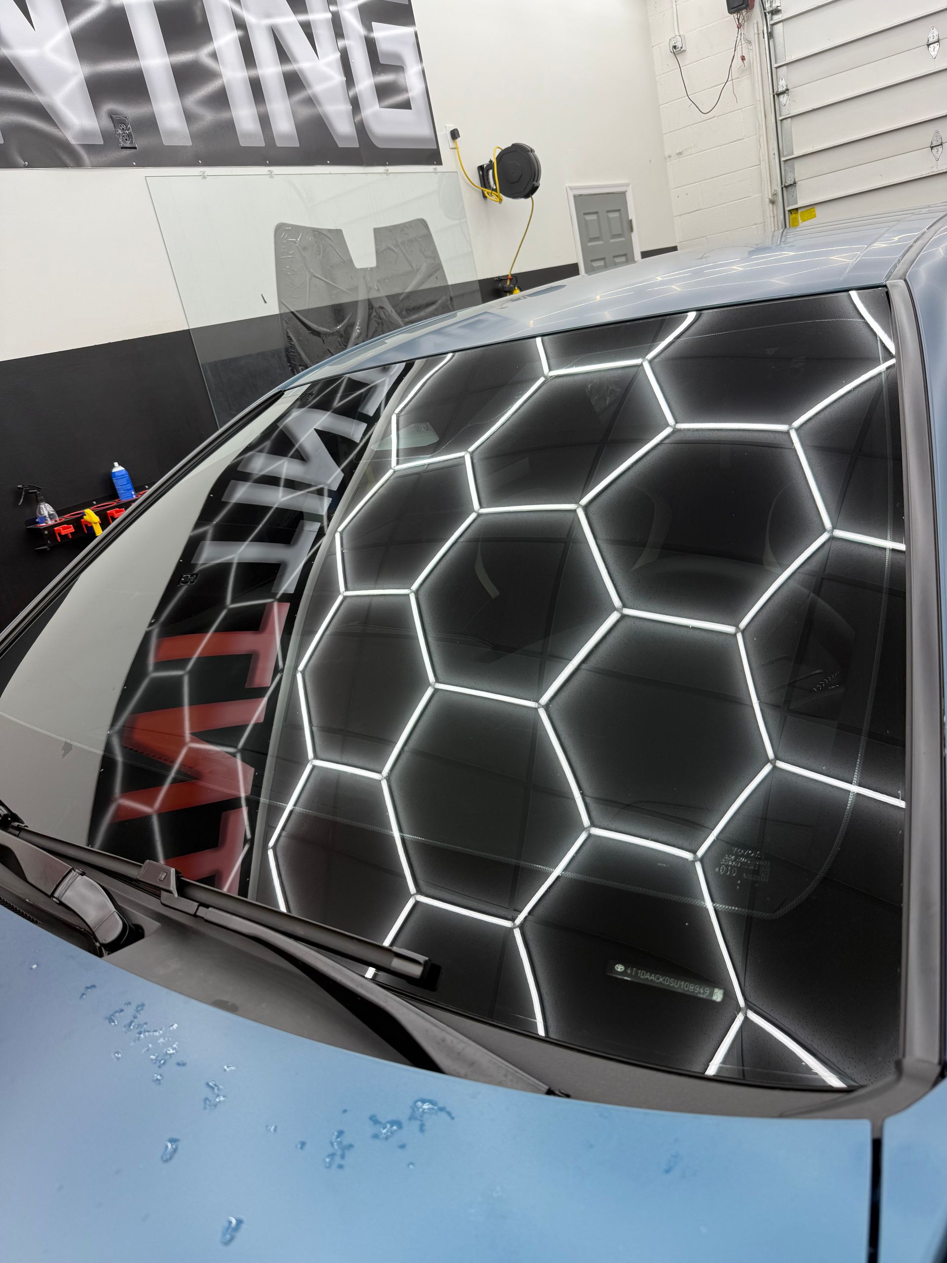 A car's windshield reflecting a honeycomb-patterned light fixture, in a detailing shop.