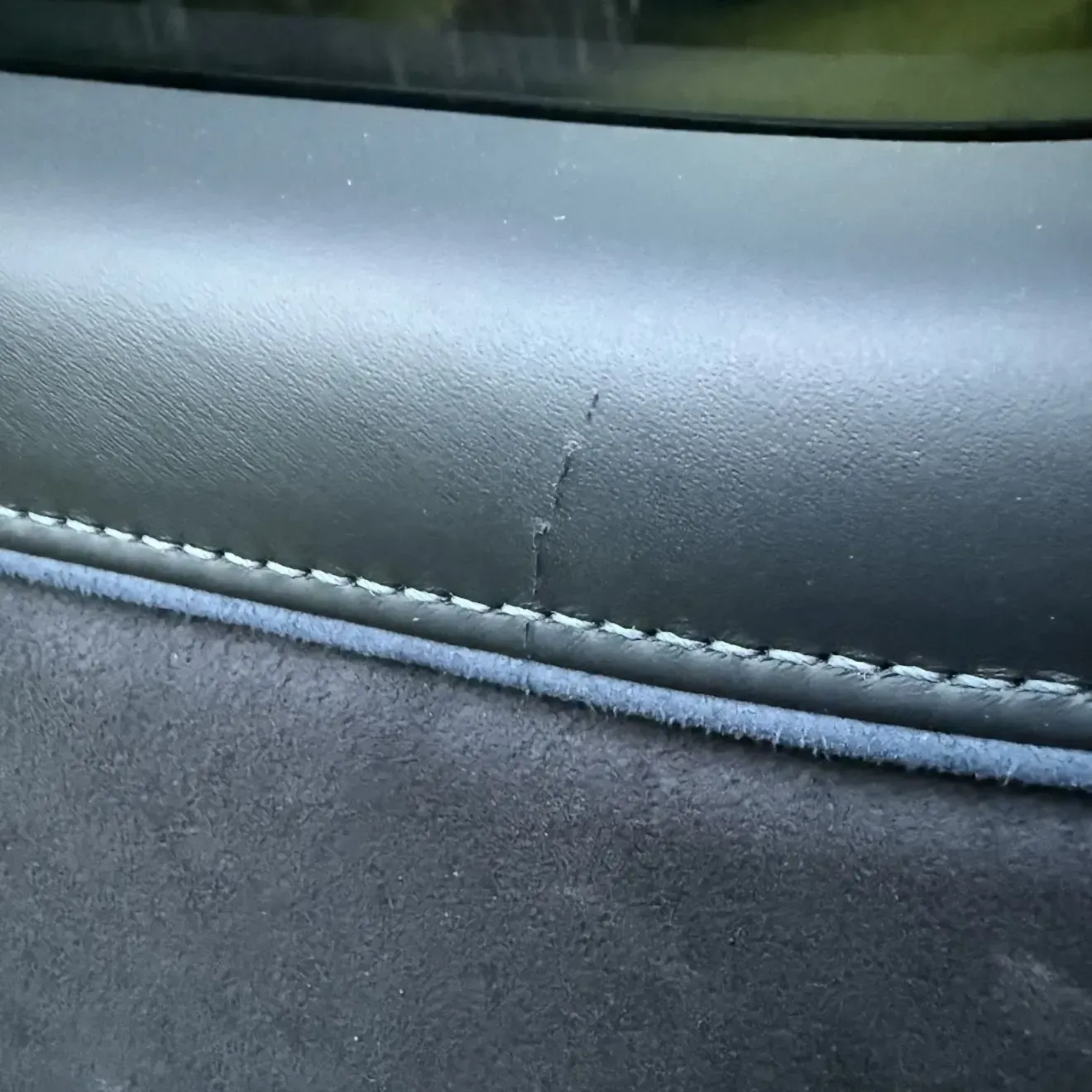 Close-up of a car door panel showing a crack in the black leather trim, with contrasting white stitching.