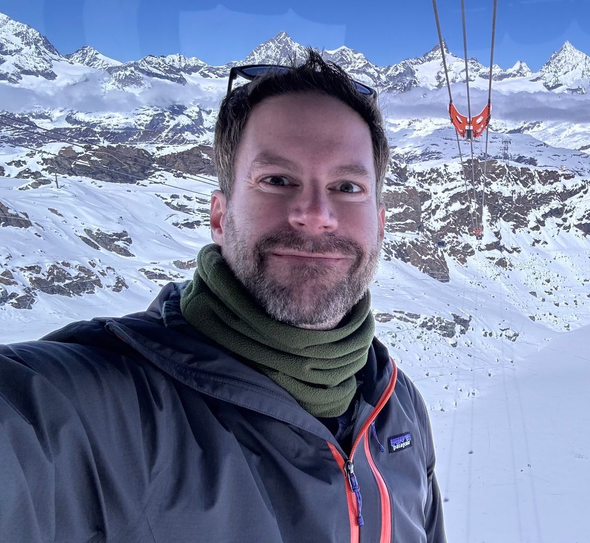 A man with a beard is taking a selfie in front of a snowy mountain.