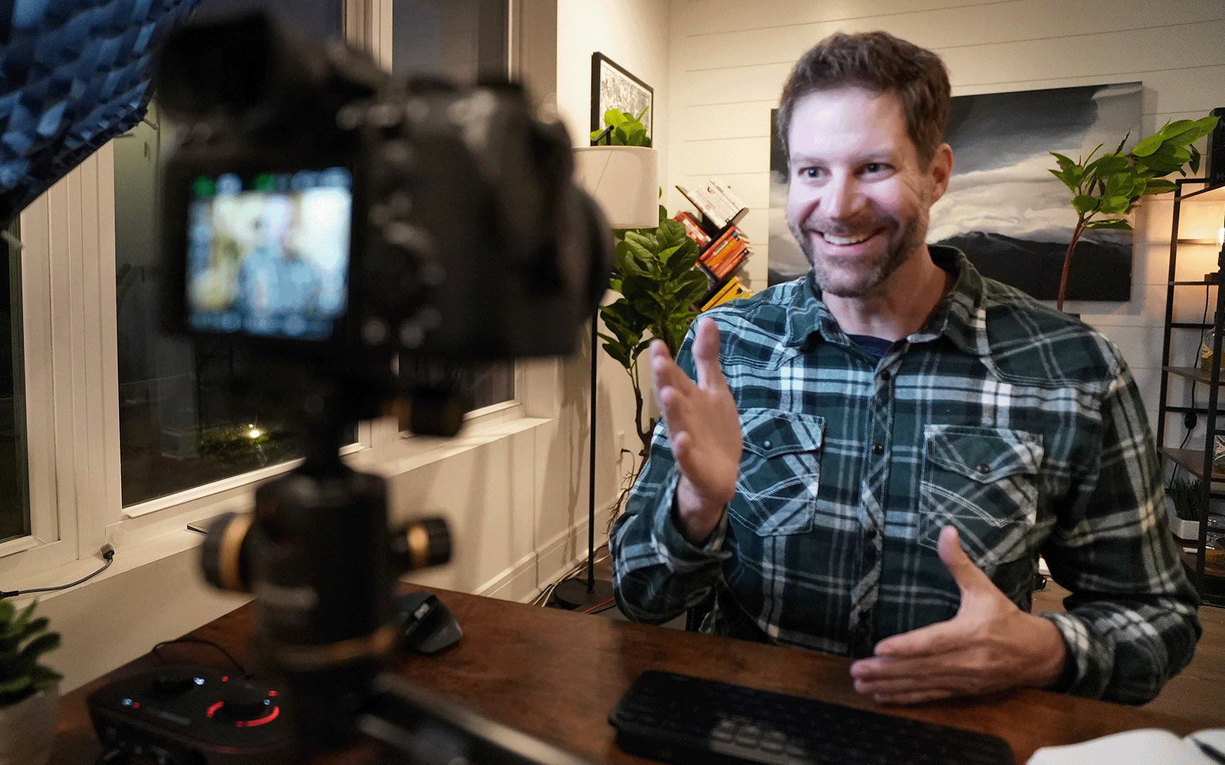 A man in a plaid shirt is sitting at a desk in front of a camera.