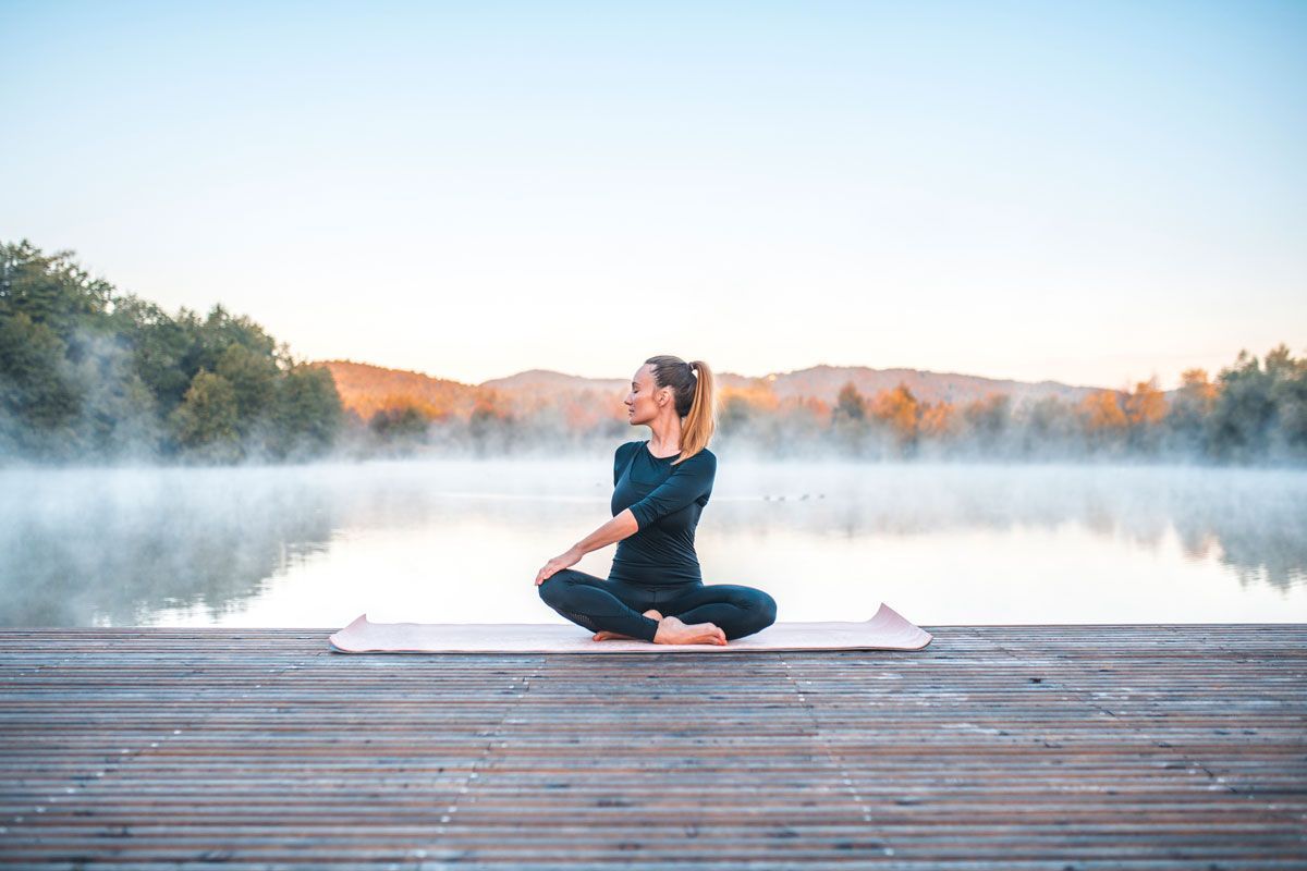 A woman is sitting on a yoga mat on a dock near a lake.