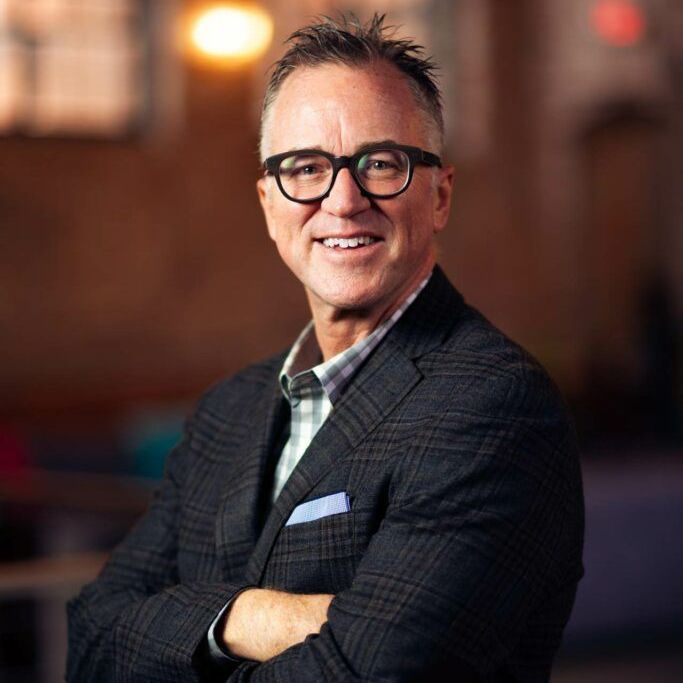 A man in a suit and glasses is standing with his arms crossed and smiling.