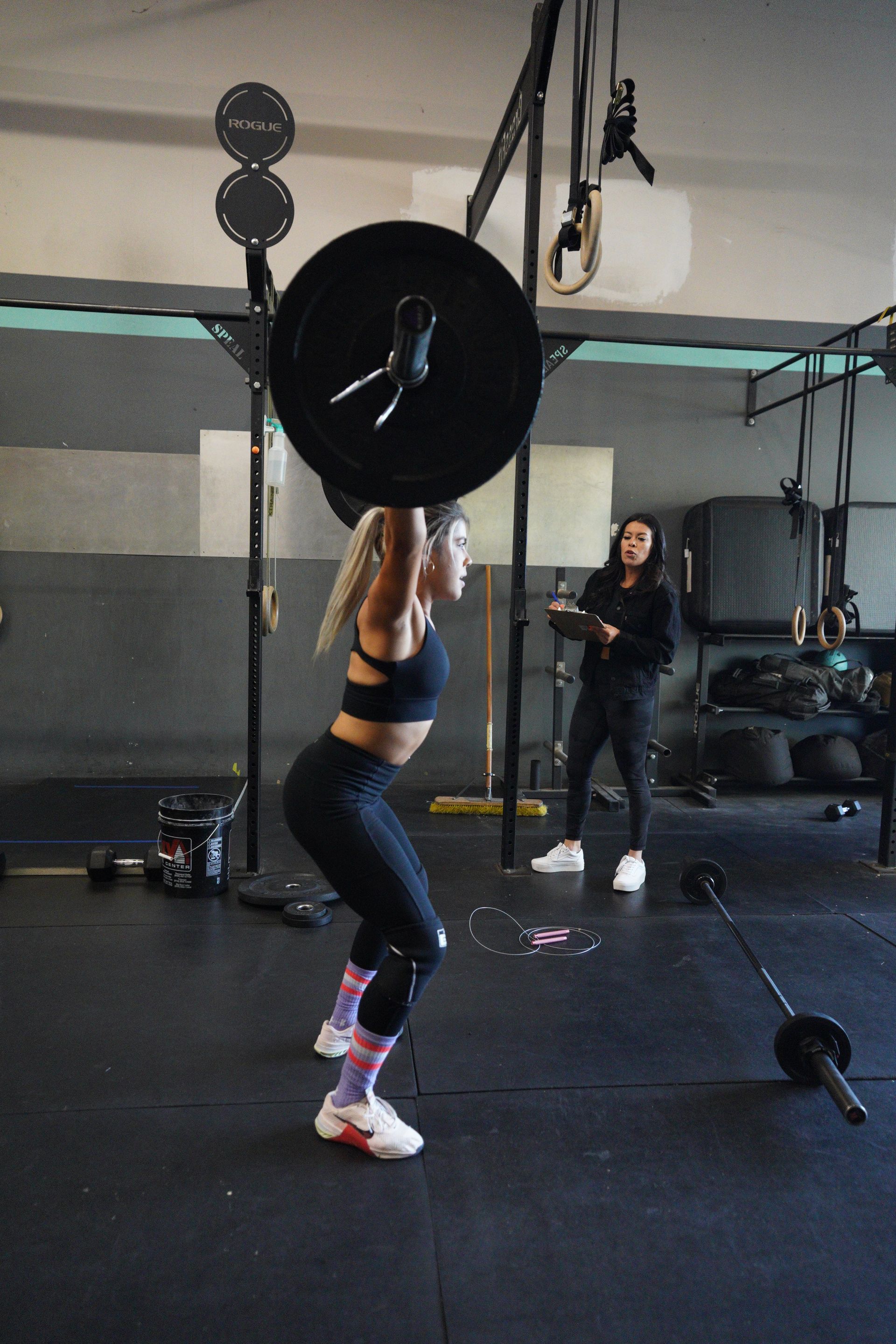 Woman in workout gear presses barbell overhead in a gym. Trainer watches.
