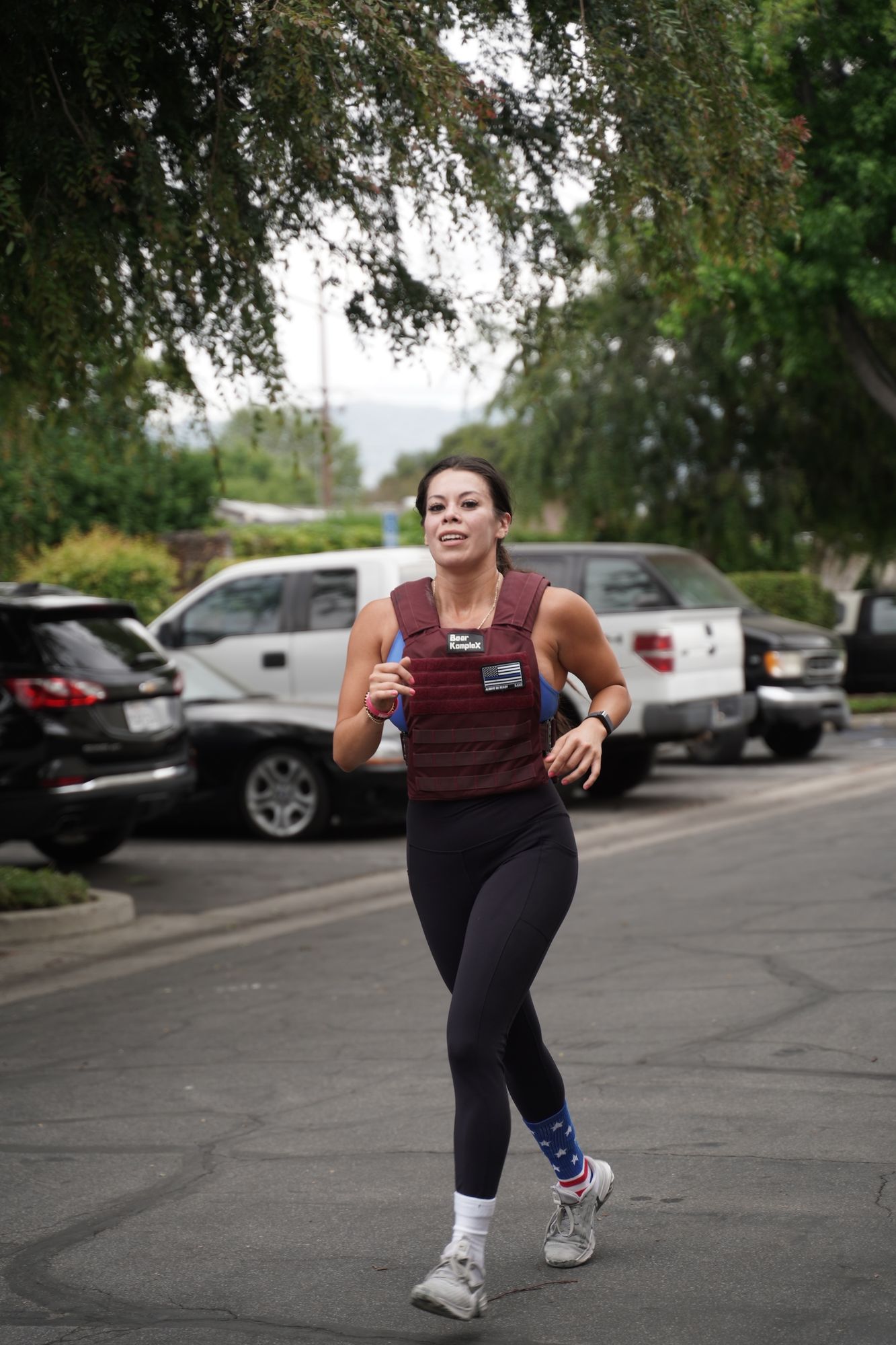 Woman runs on street wearing weighted vest, black leggings, and patriotic socks. Cars in the background.