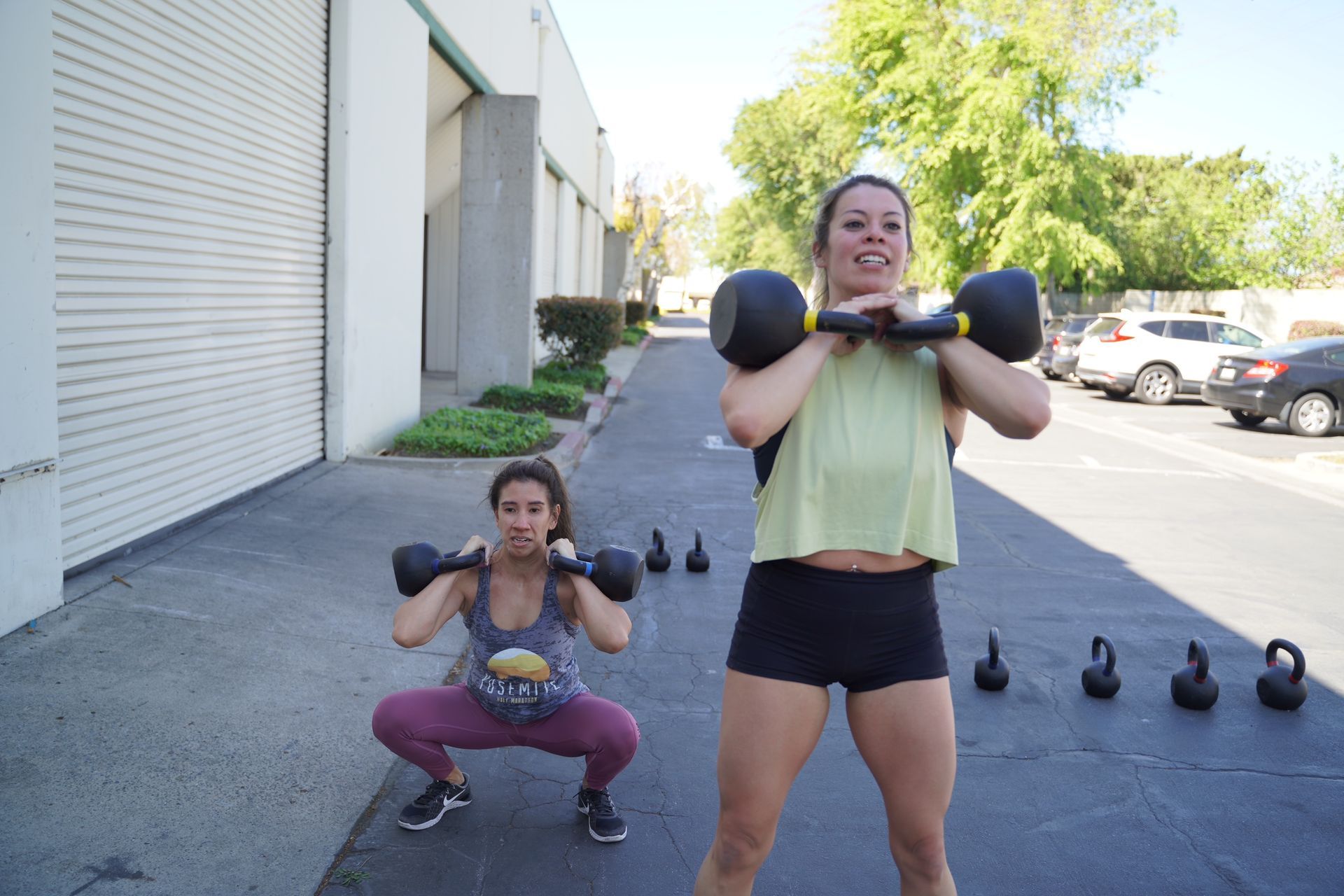 Two women exercising with kettlebells outdoors: one squatting, one standing.